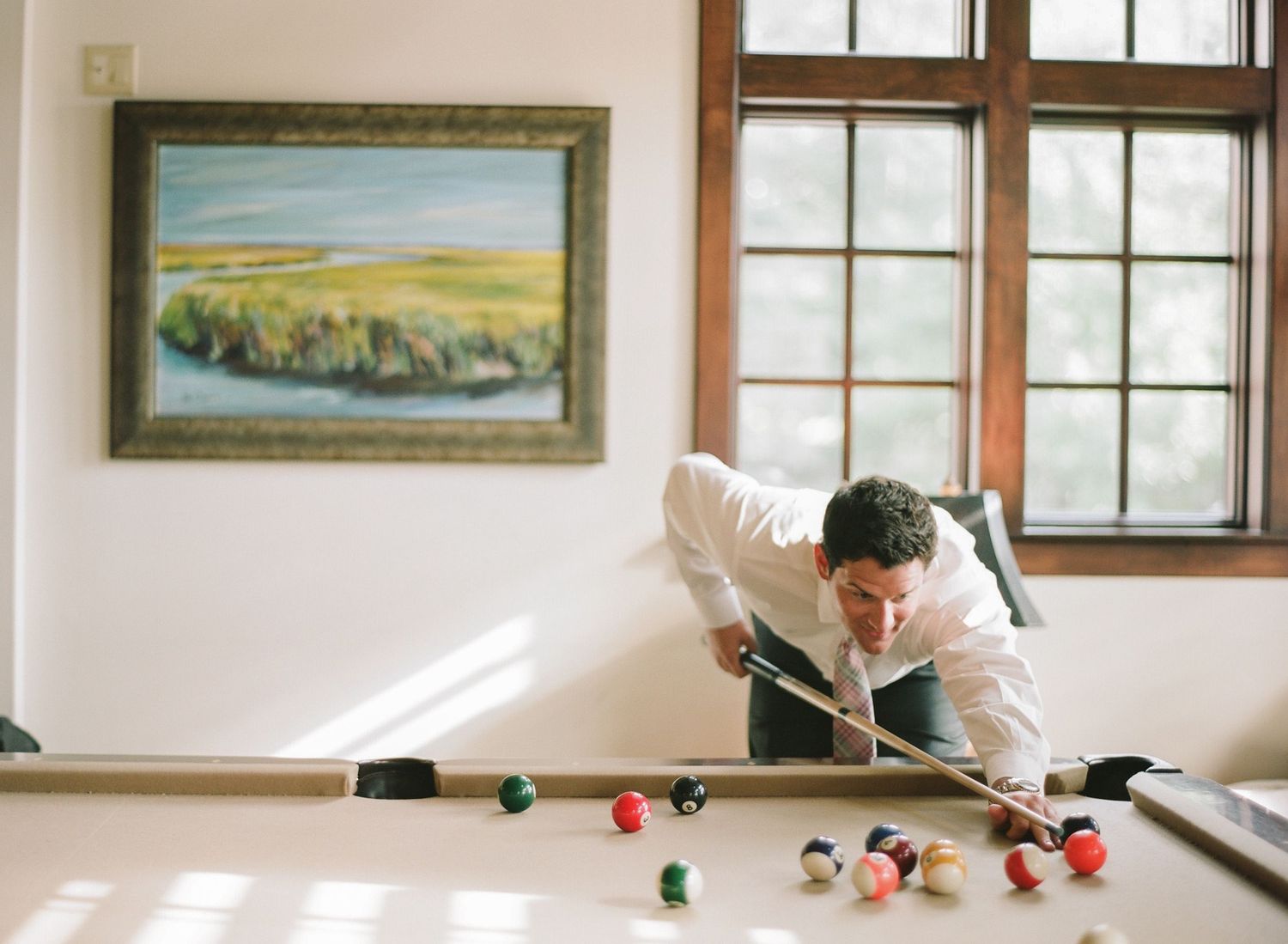 Person leaning over pool table in sunlit room with landscape paintings on walls.