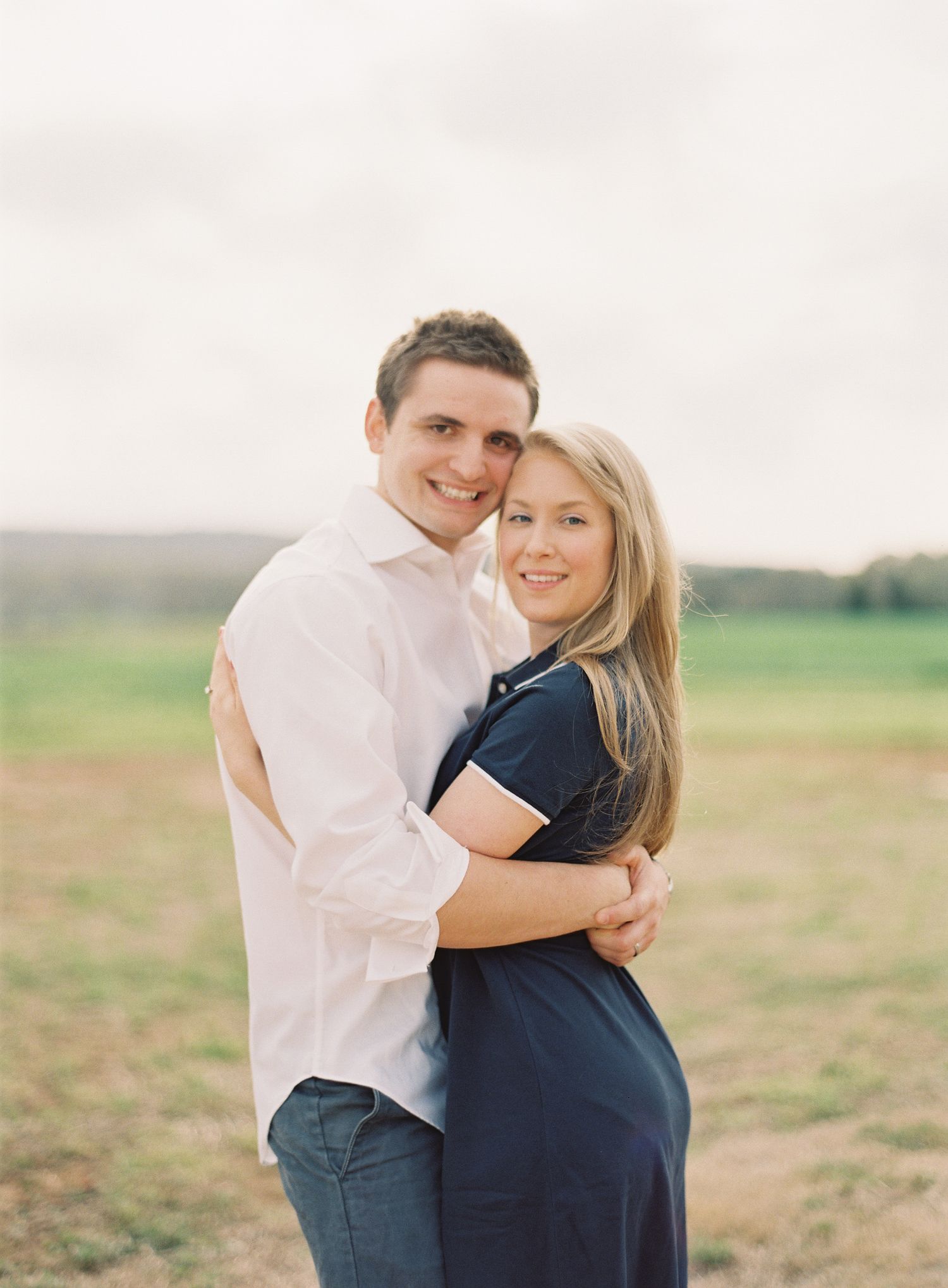 A couple embraces lovingly in a rustic outdoor setting with rolling hills in the background.