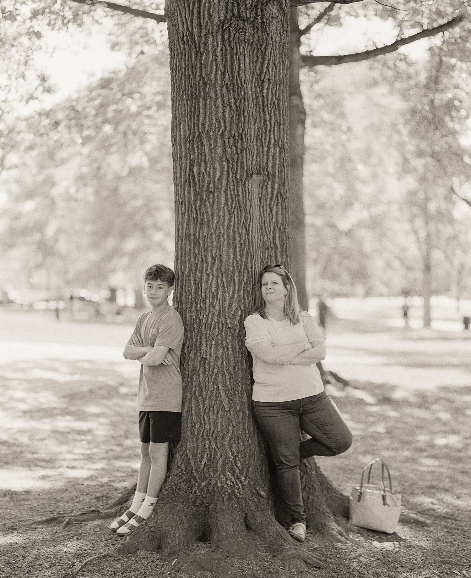A boy and woman lean against a large tree in a park, arms crossed, in a black and white photo.