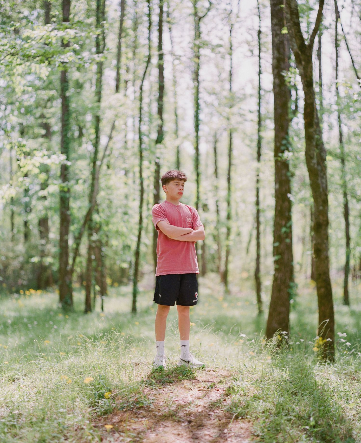 Young man in red shirt and black shorts standing with arms crossed on a forest path among tall green trees.