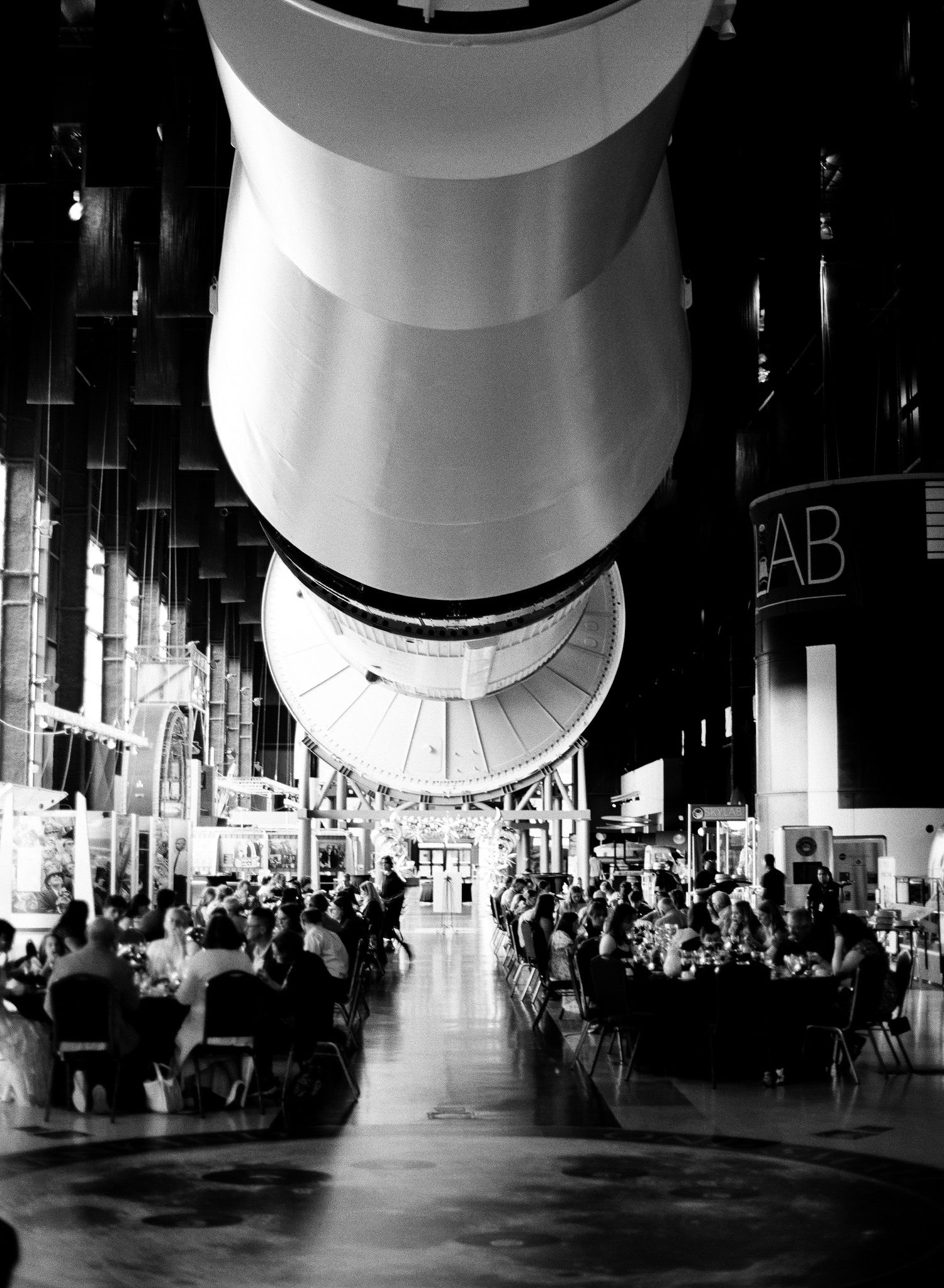 Black and white photo of guests dining beneath a massive rocket engine at a space museum event.