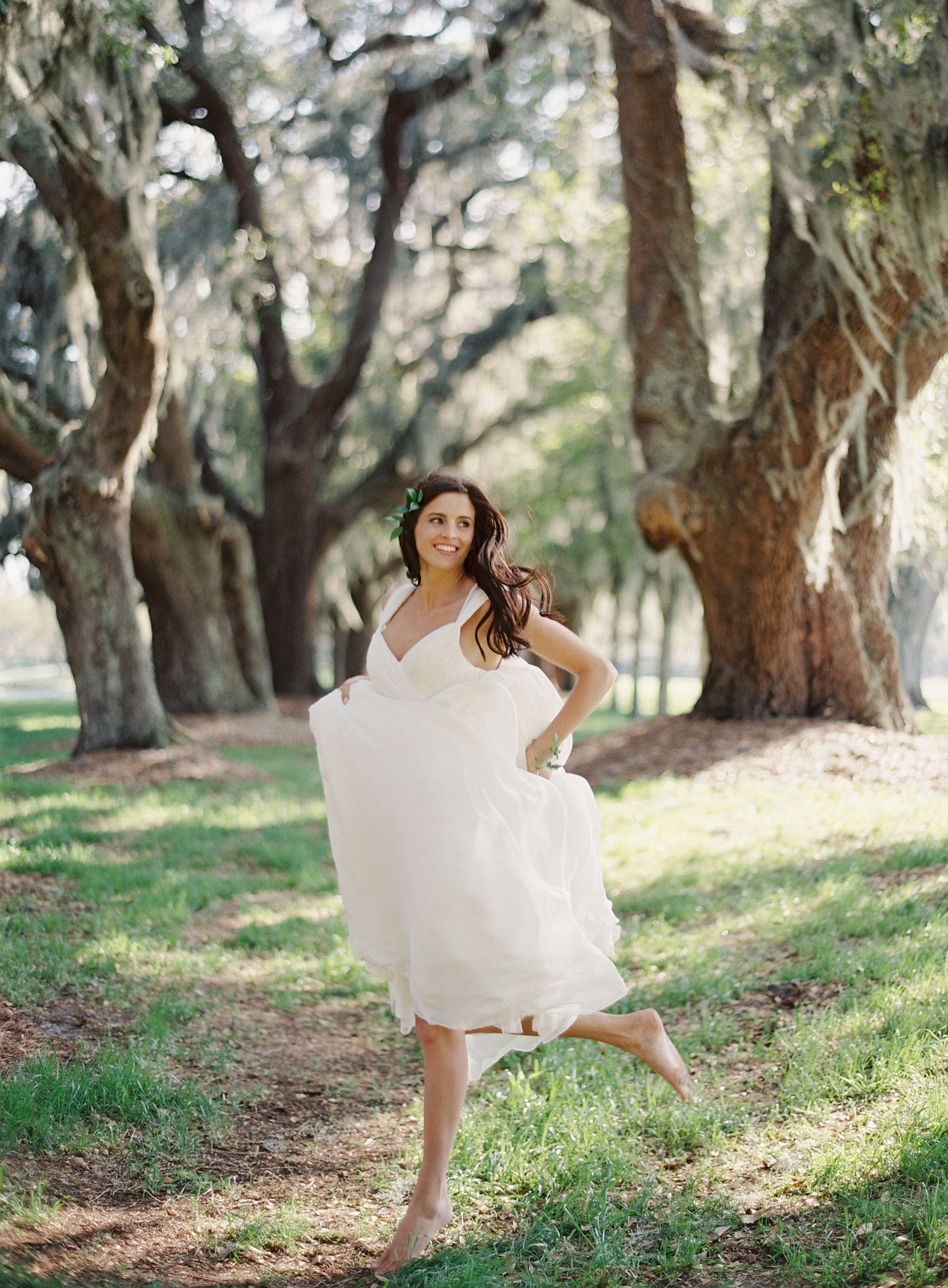 Bride in white dress twirling playfully under a canopy of oak trees.