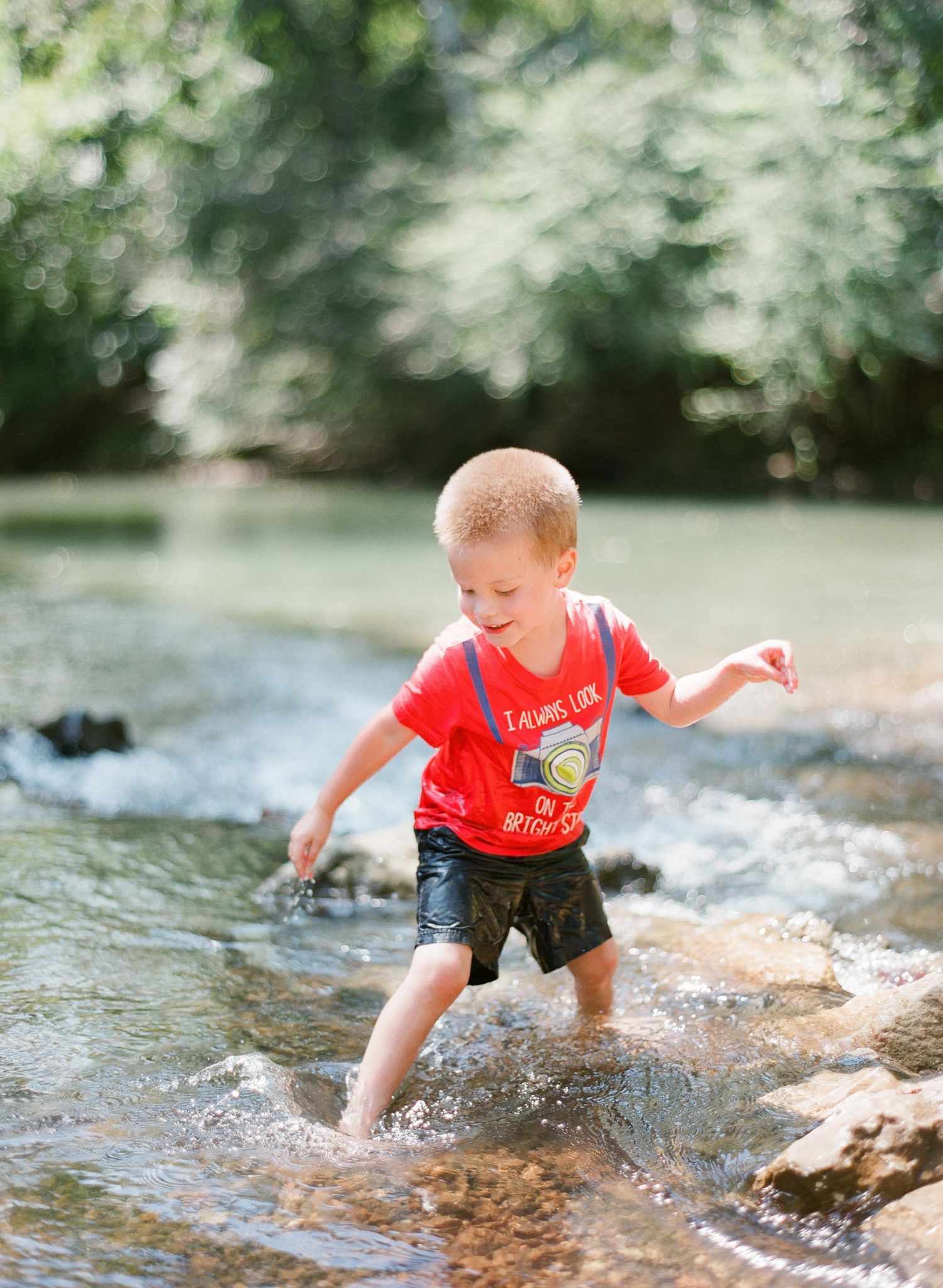 Child in bright red shirt playing and jumping in a shallow woodland stream.