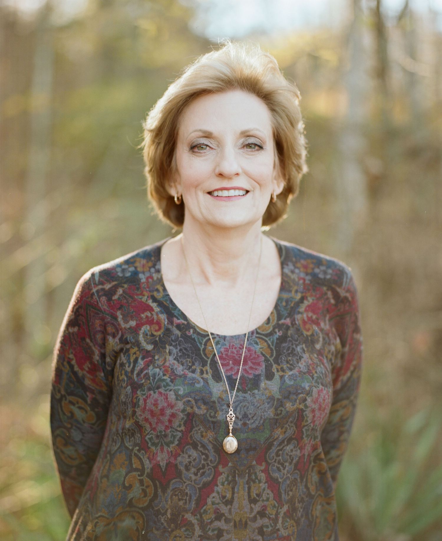 Woman in floral print dress smiling warmly against autumn forest backdrop.