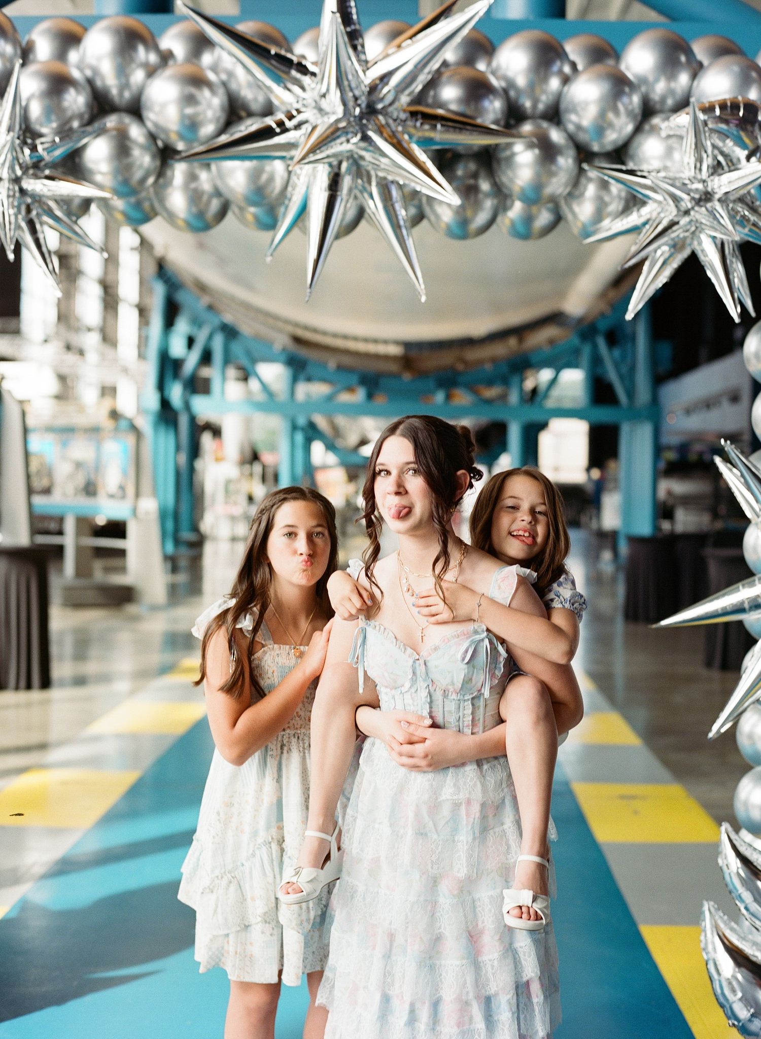 Three girls pose under silver star and balloon decorations in a bright indoor venue, wearing matching light blue dresses.