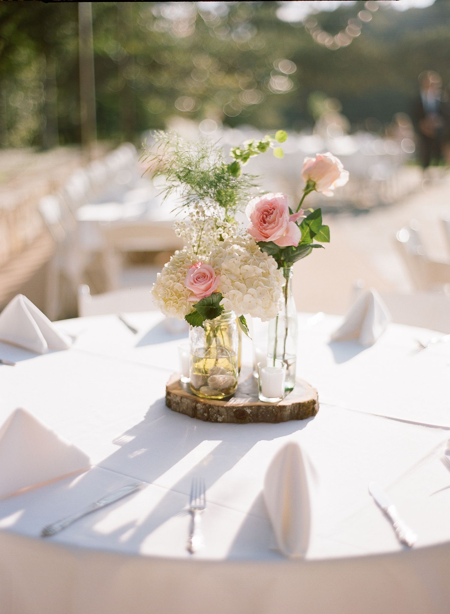 Elegant table centerpieces with pink roses and white flowers in glass vases on white tablecloths.