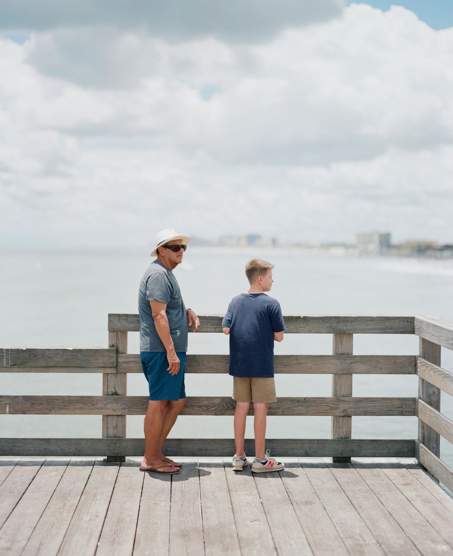 Two figures stand at the railing of a wooden pier overlooking the ocean.