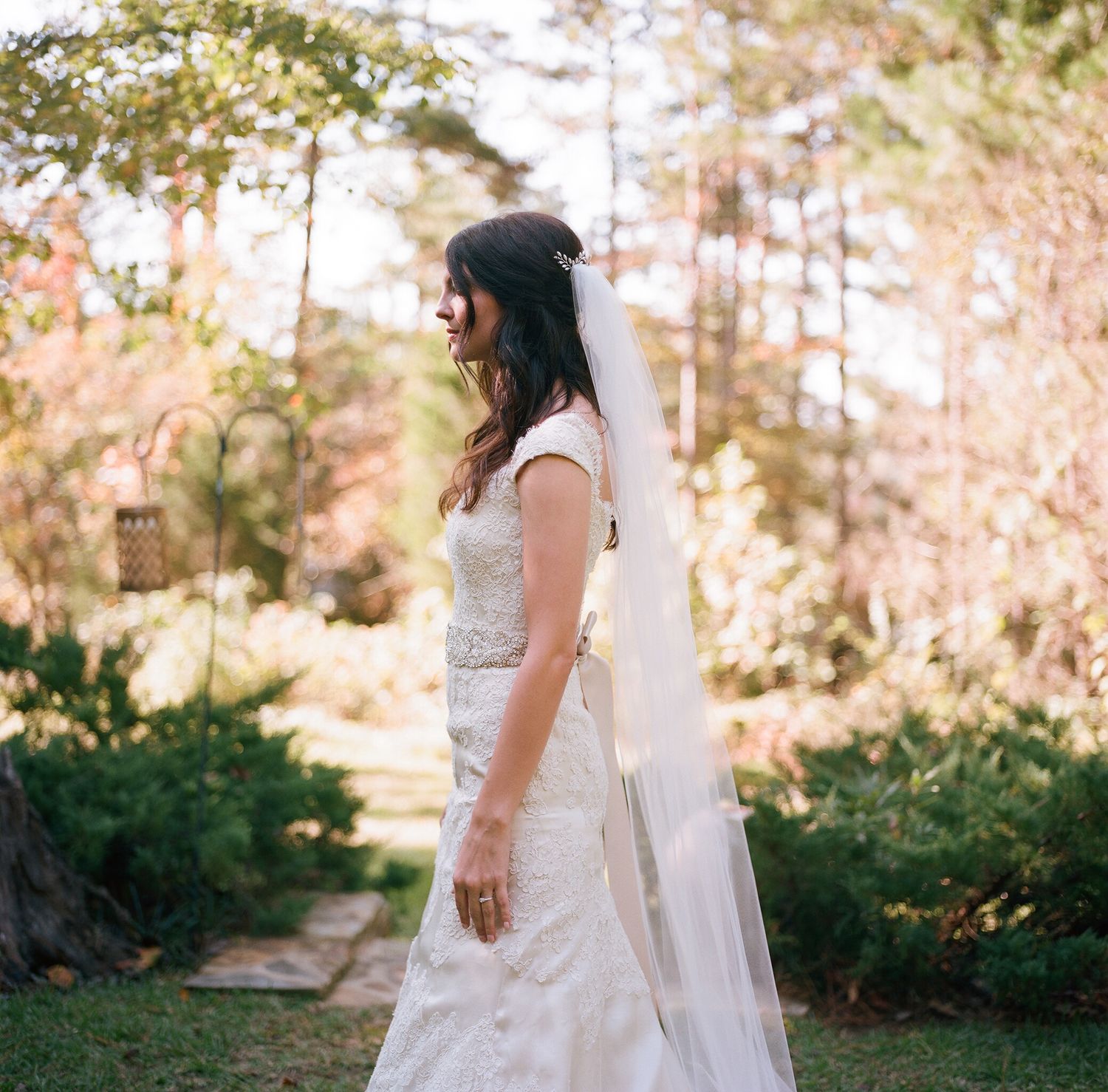 Bride in flowing white gown and veil photographed in sunlit outdoor garden setting.