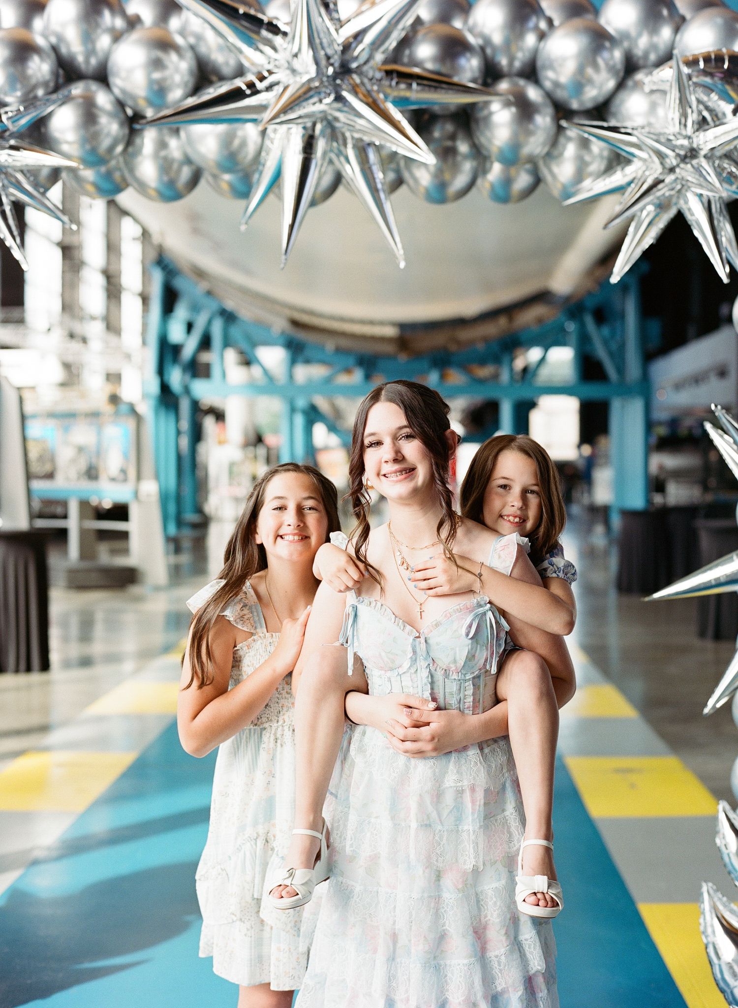 Three smiling girls in light dresses pose together under silver star balloons in a colorful event venue.