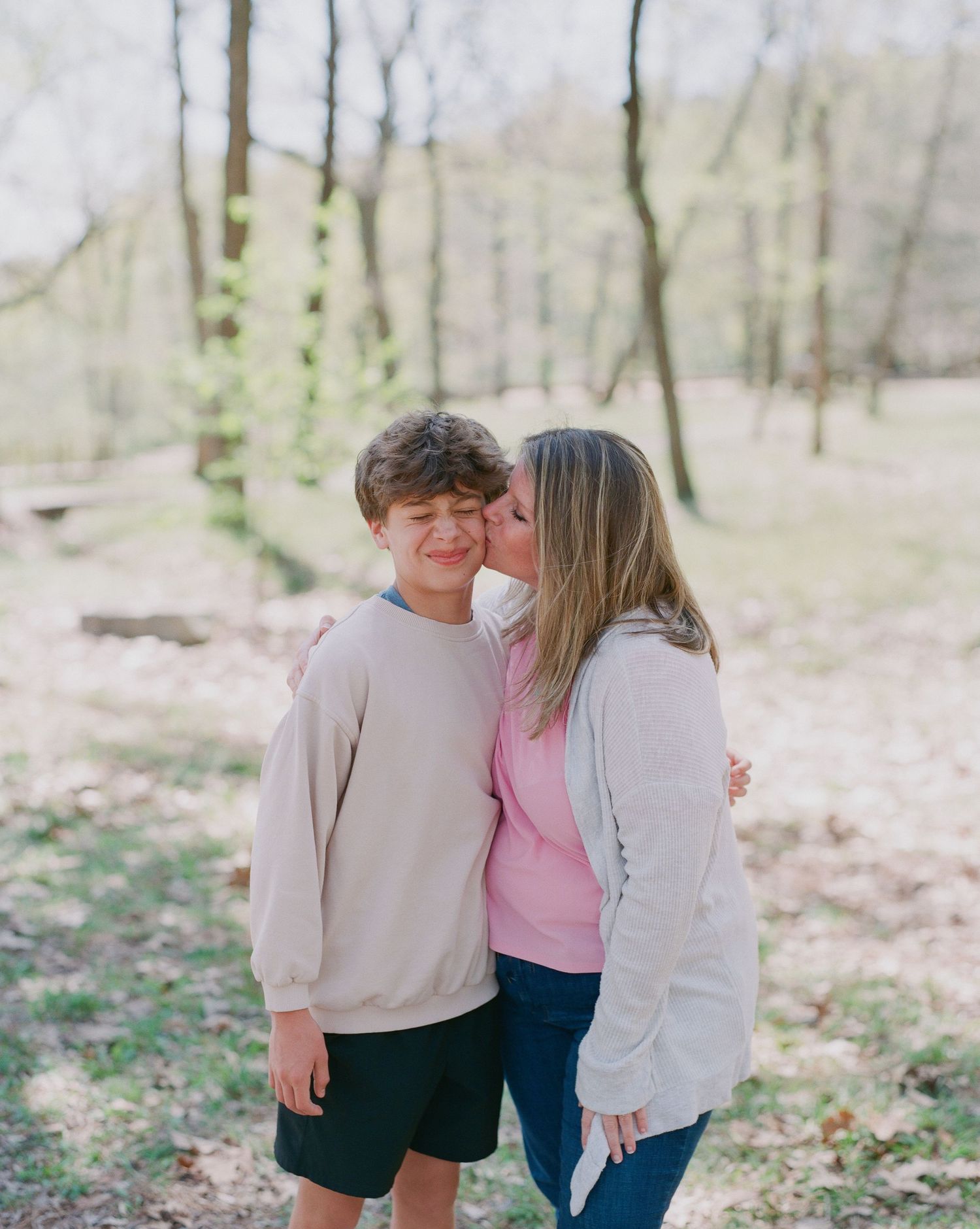 A woman kisses a smiling boy on the cheek outdoors in a wooded park setting during spring.