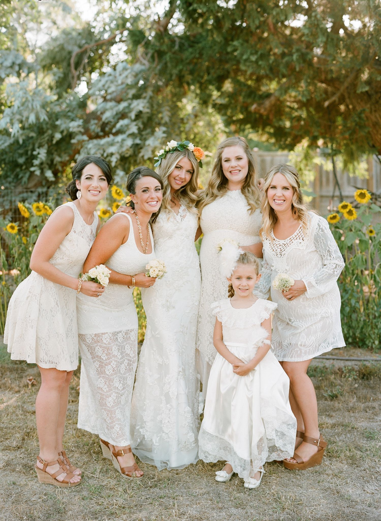Bridesmaids in white lace dresses pose together outdoors in a garden setting.