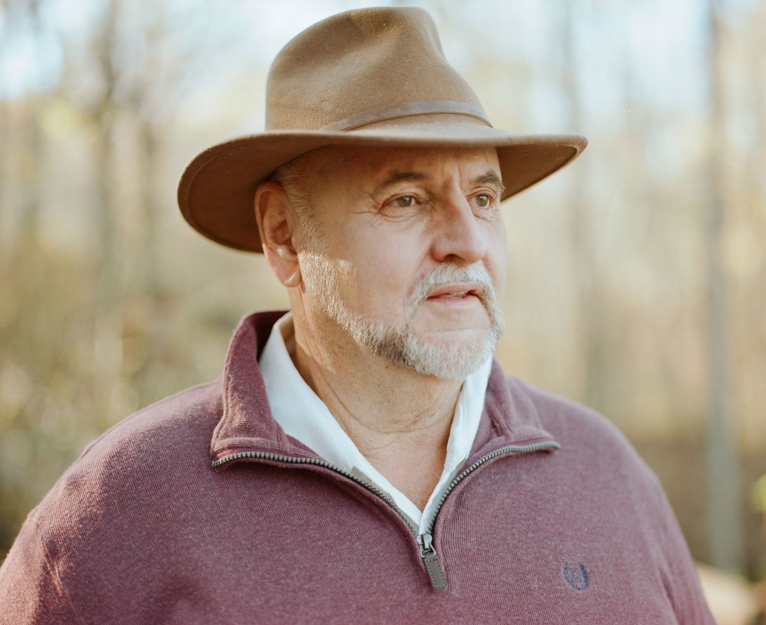 Portrait of a man wearing a brown hat and burgundy jacket in an autumn forest setting.