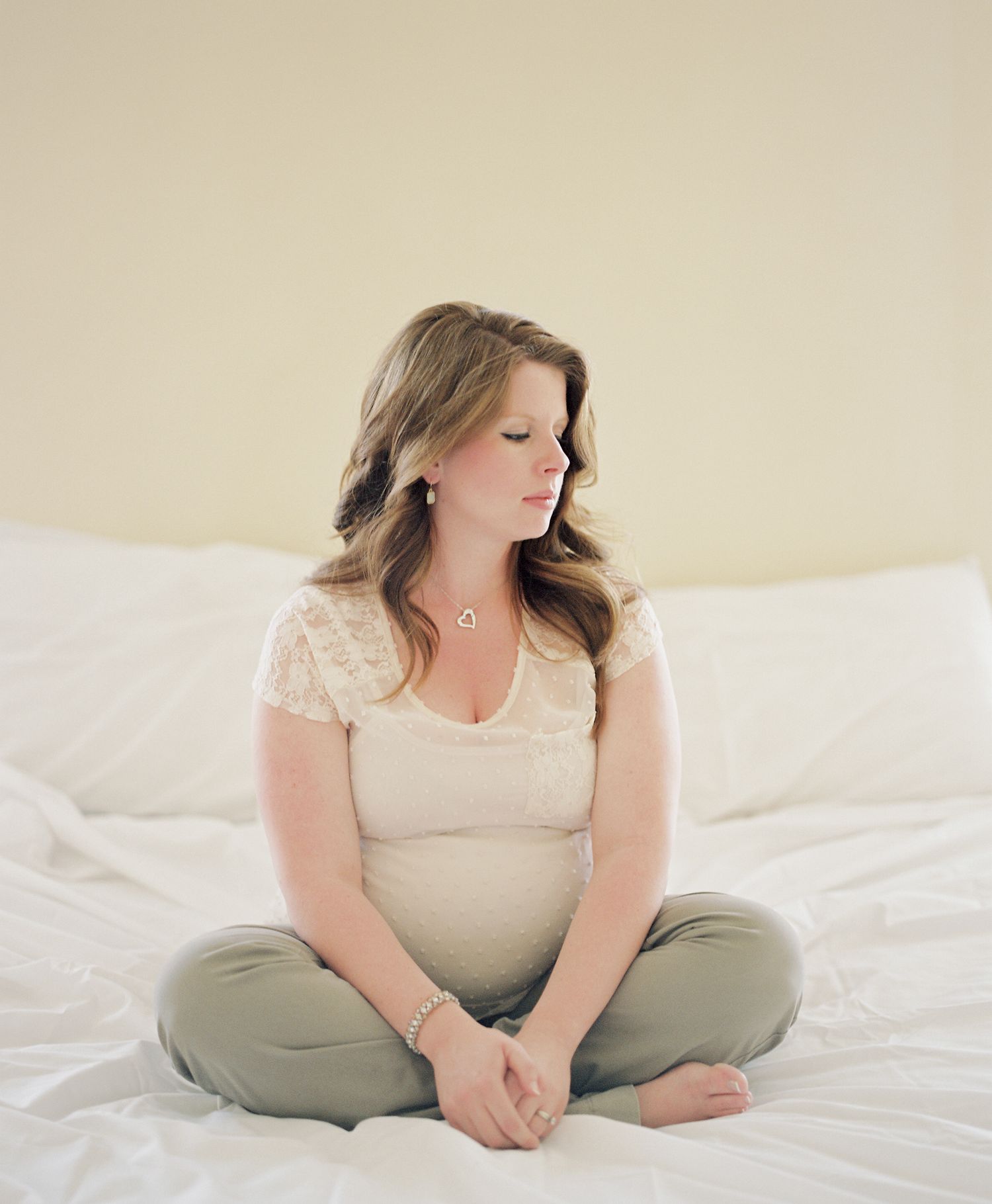Expectant mother in white top sitting cross-legged on white bedding.
