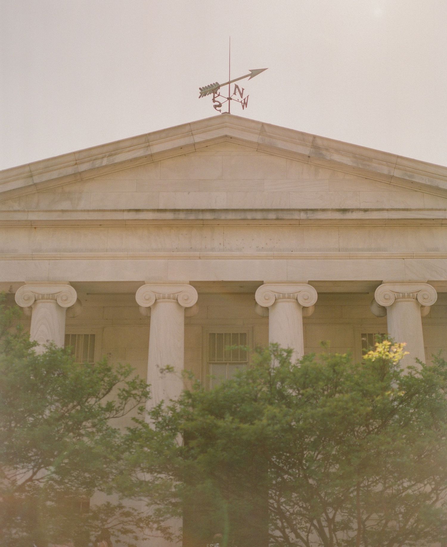 Neoclassical building facade with Ionic columns, triangular pediment, and weather vane surrounded by lush green trees.