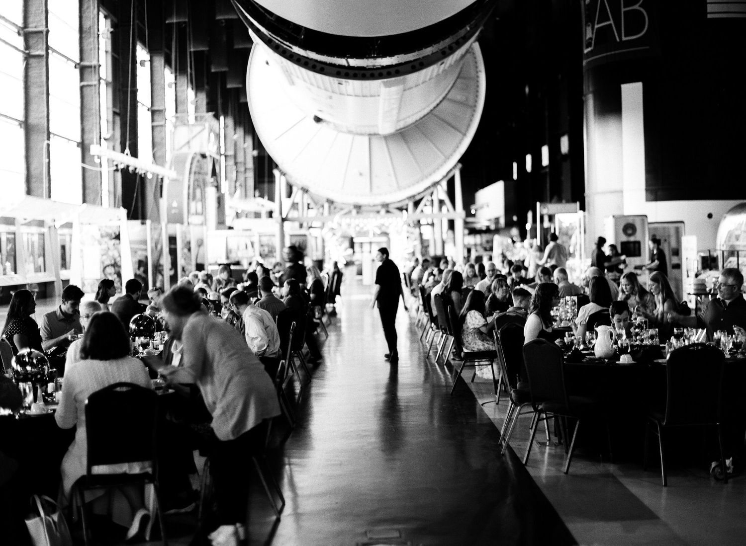 Black and white photo of an elegant event under a large rocket in a museum hall with guests dining at long tables.