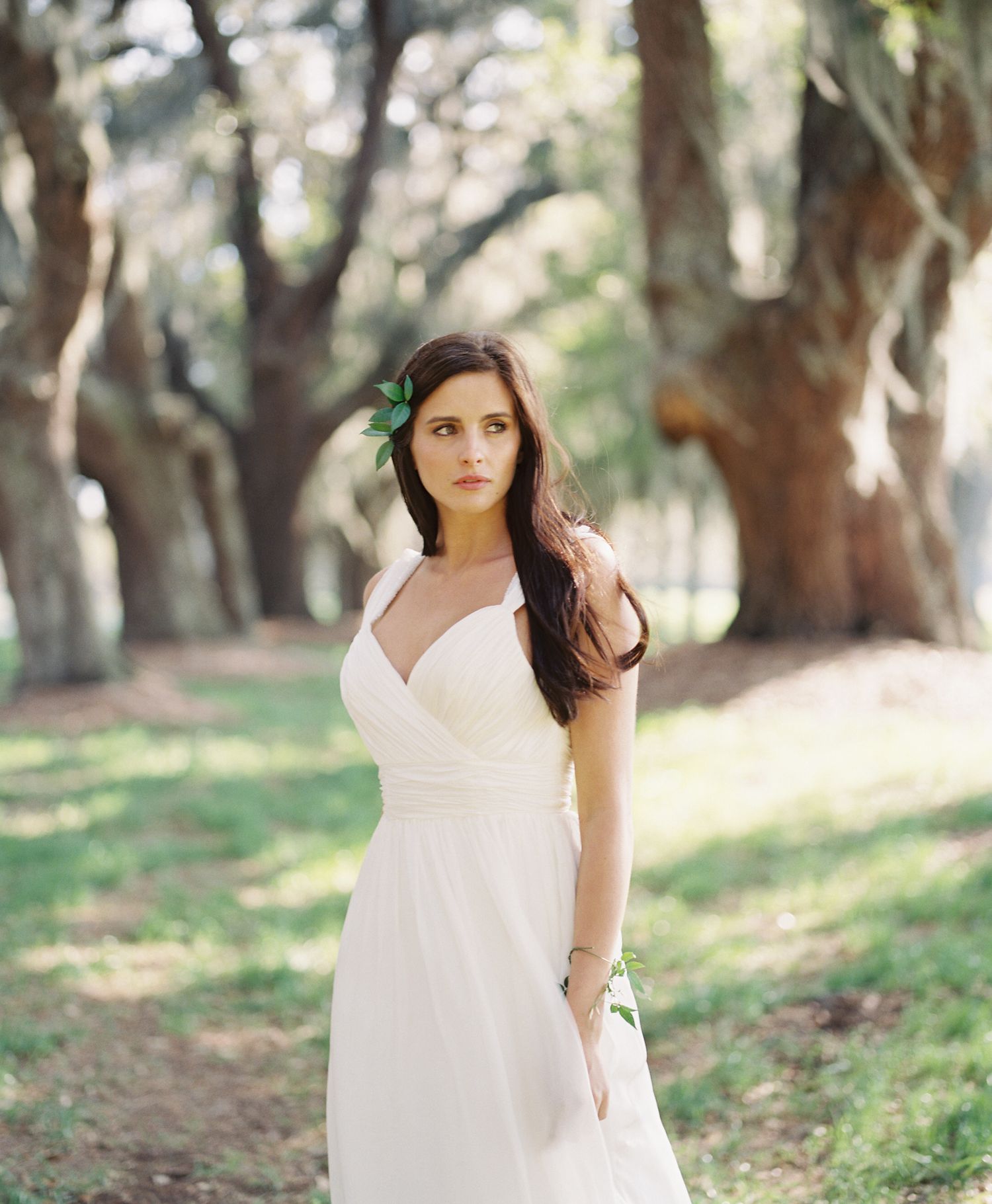 Elegant bride in white dress poses in a dreamy outdoor garden setting with sunlit trees in background.
