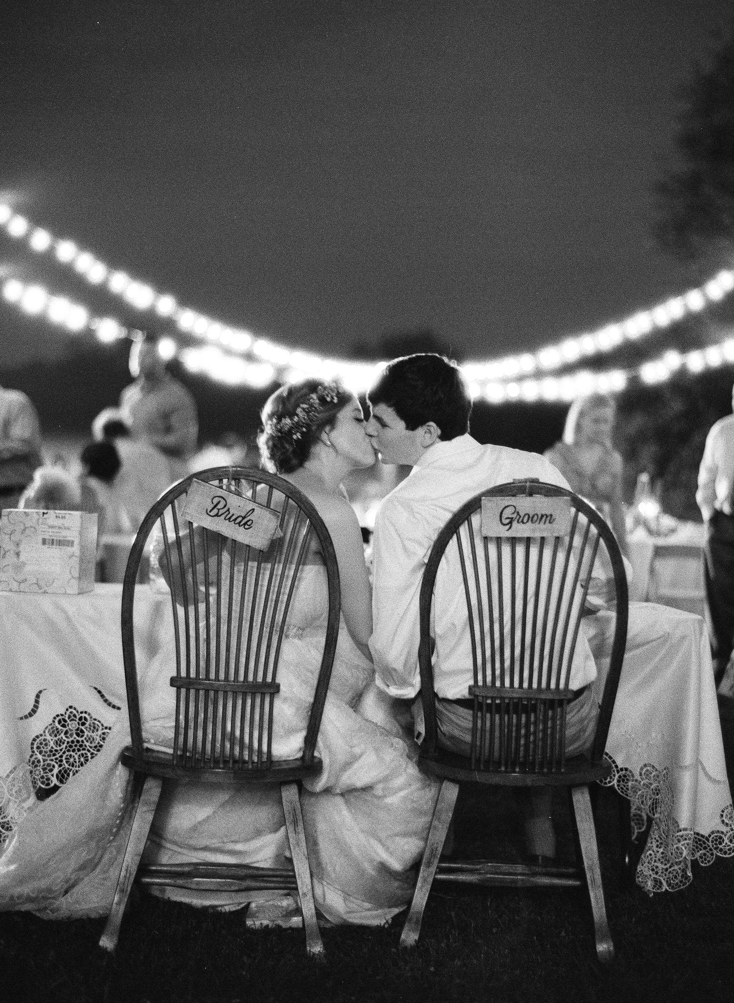 Row of wooden chairs with guests at outdoor evening wedding reception under string lights.