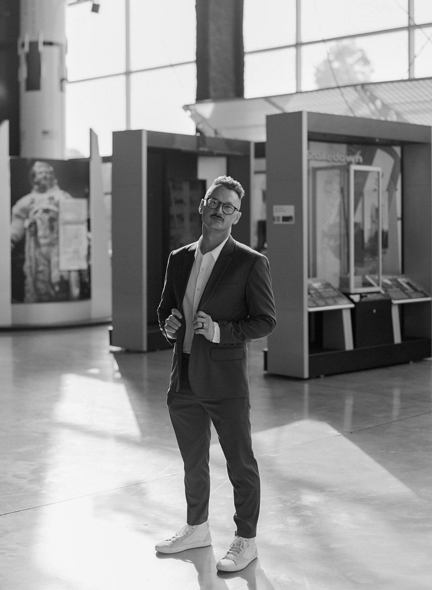 Man in suit and white sneakers poses in modern museum lobby, black and white photo.
