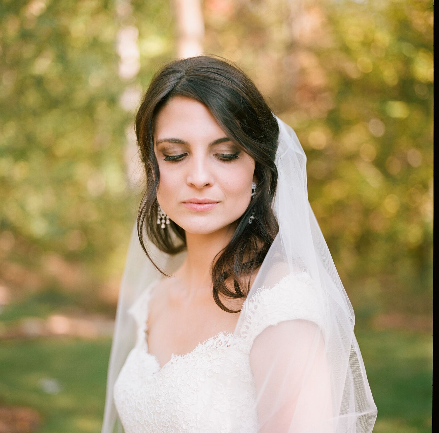 Bride wearing delicate veil and lace dress poses in natural outdoor setting.