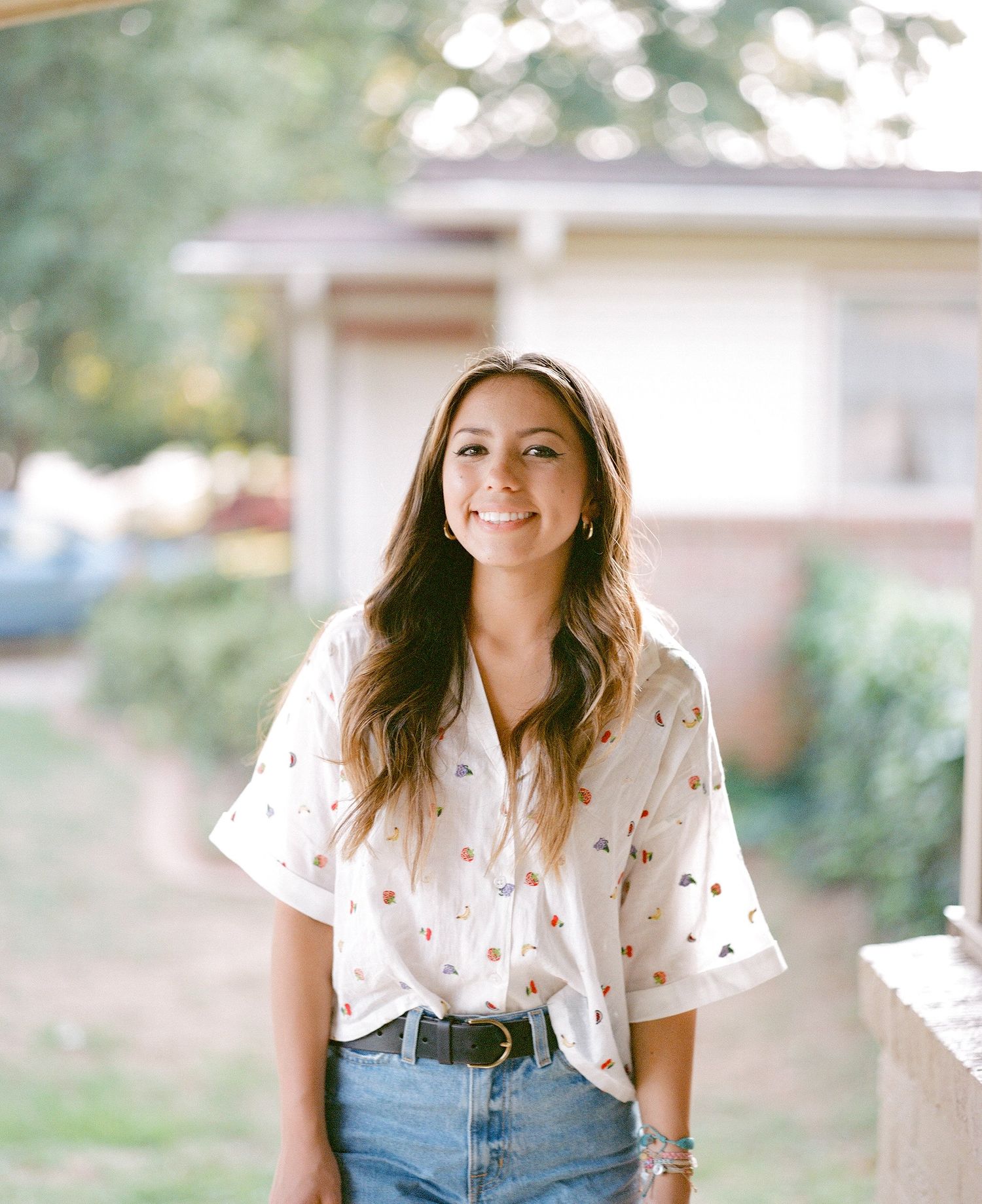 A person in a white patterned shirt and jeans poses outdoors in soft natural lighting.