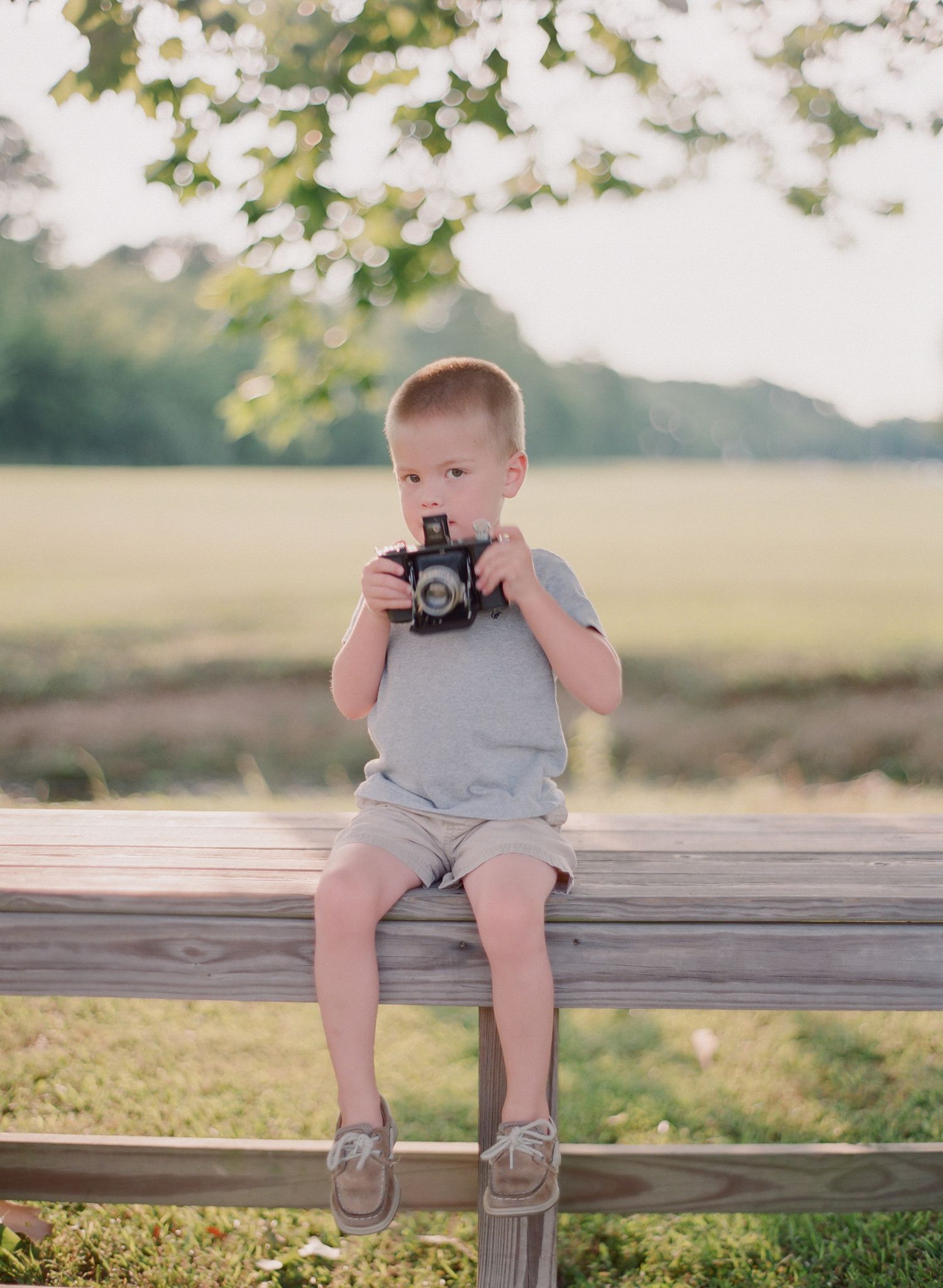 Small child sitting on wooden fence holding vintage camera in sunny field.