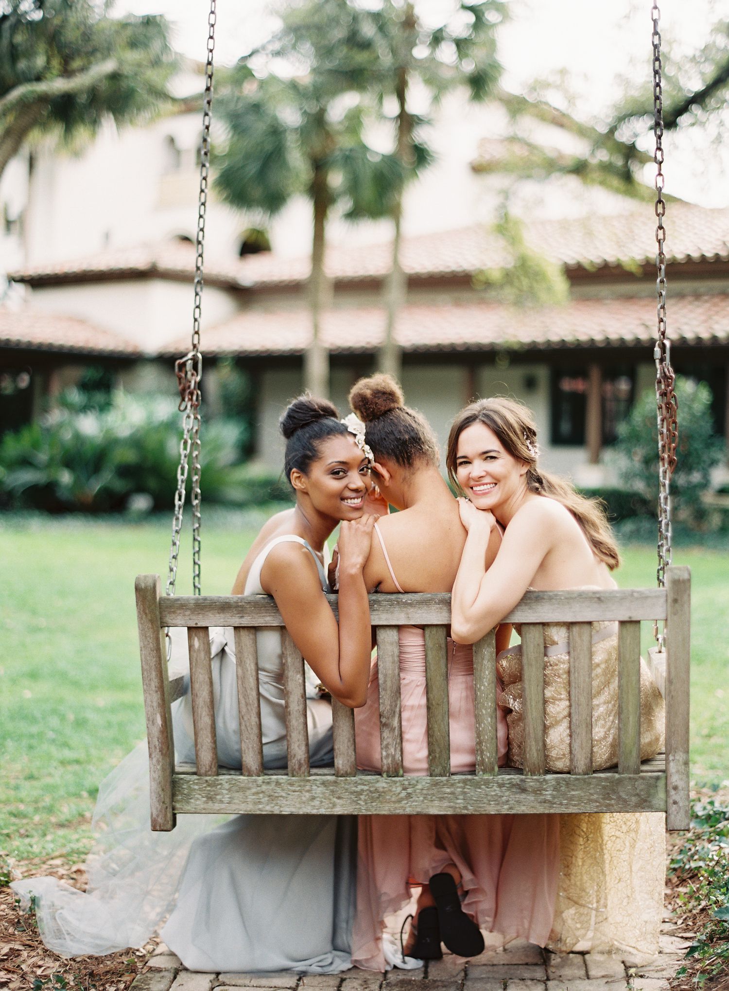 Three bridesmaids in pastel dresses share candid moment on wooden garden swing.
