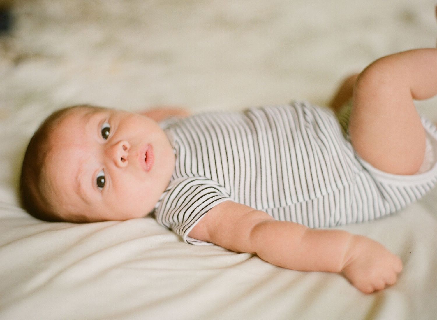 Baby in striped onesie lying on white bedding.