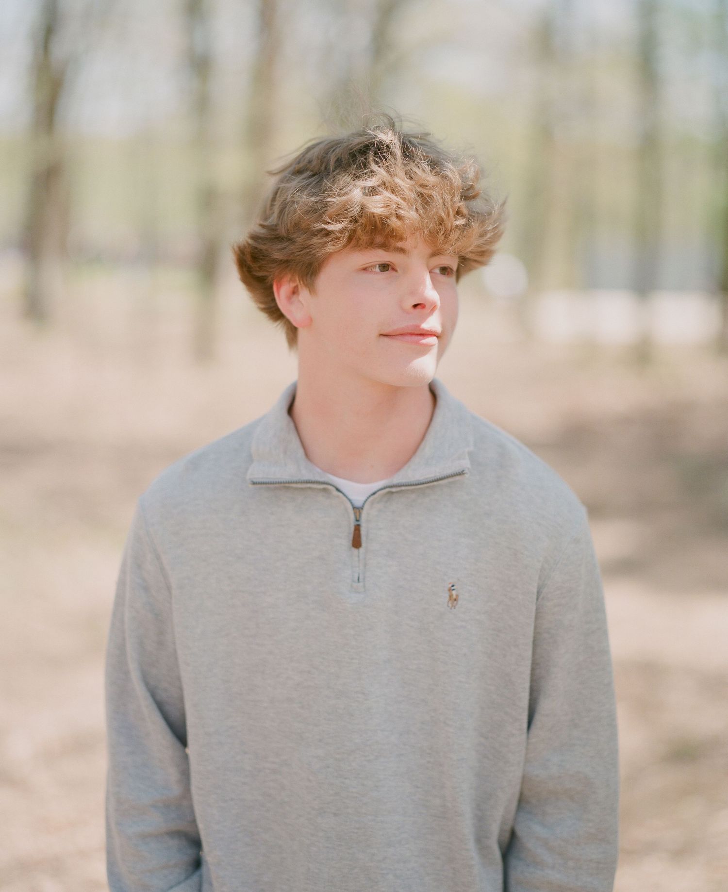 Young man with curly hair wearing a gray Polo quarter-zip sweatshirt standing outdoors among trees.