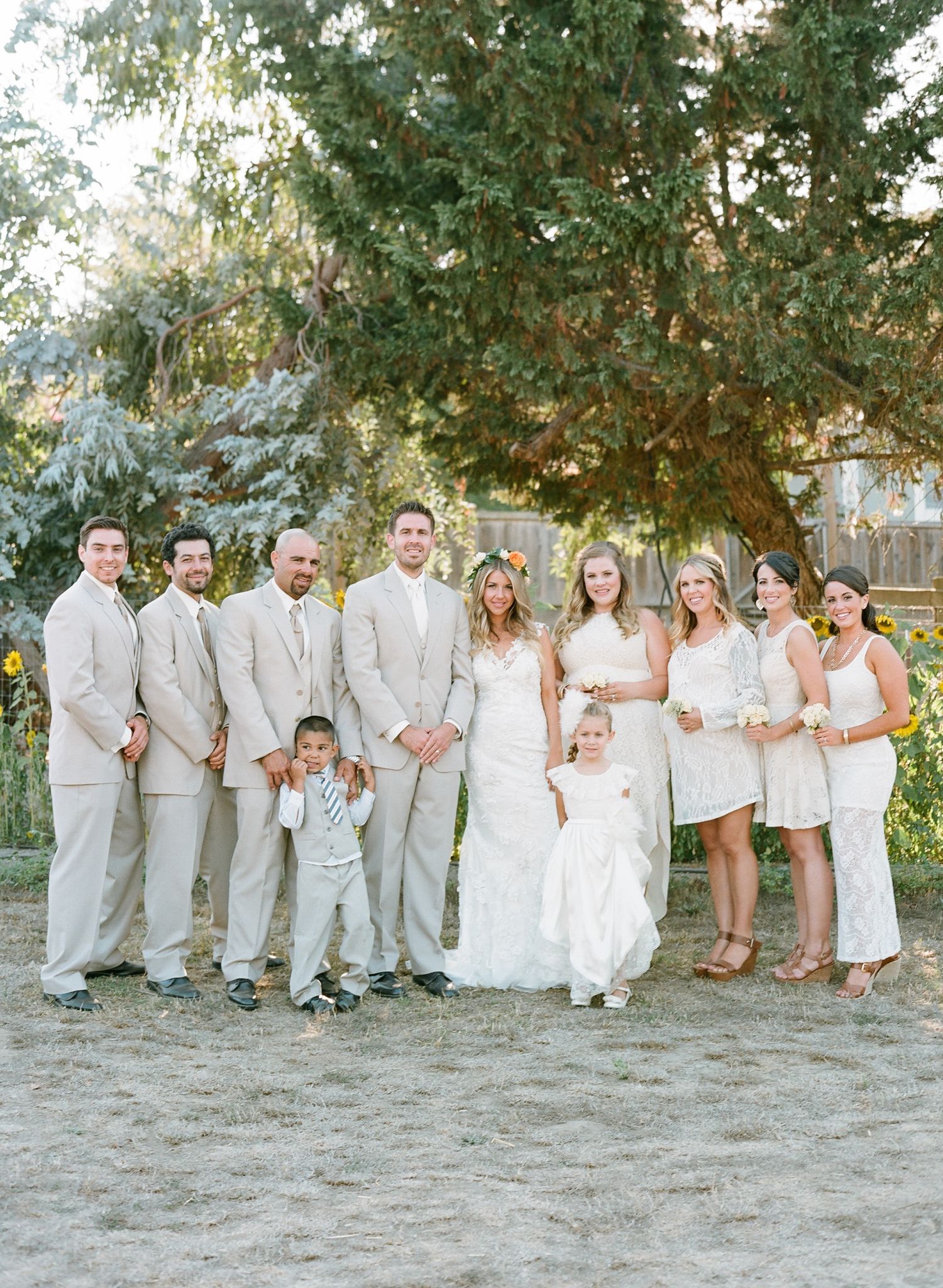 Large wedding party in white dresses and gray suits poses together under trees.