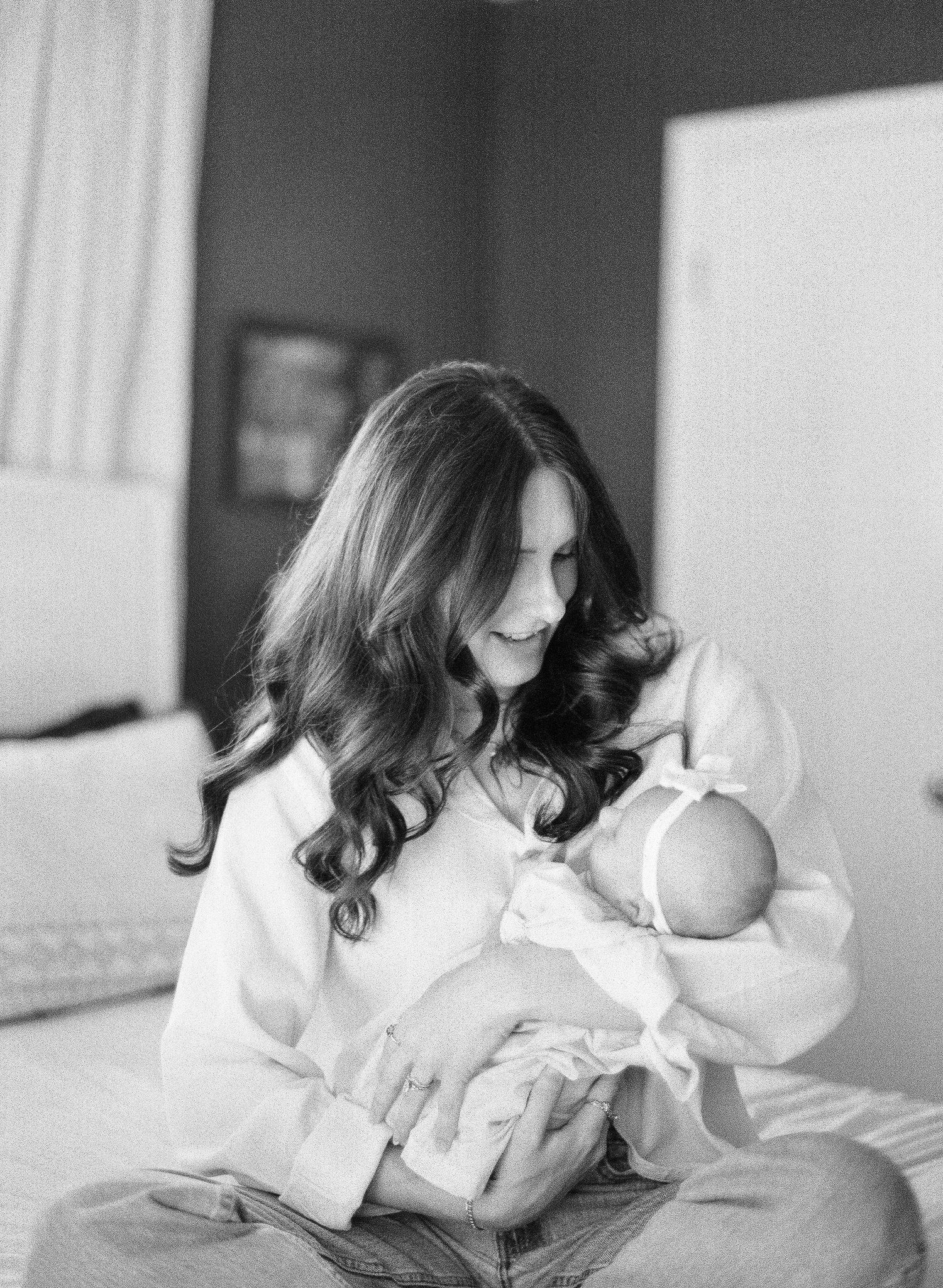 Black and white photo of a smiling mother with long wavy hair cradling her newborn baby while sitting on a bed.