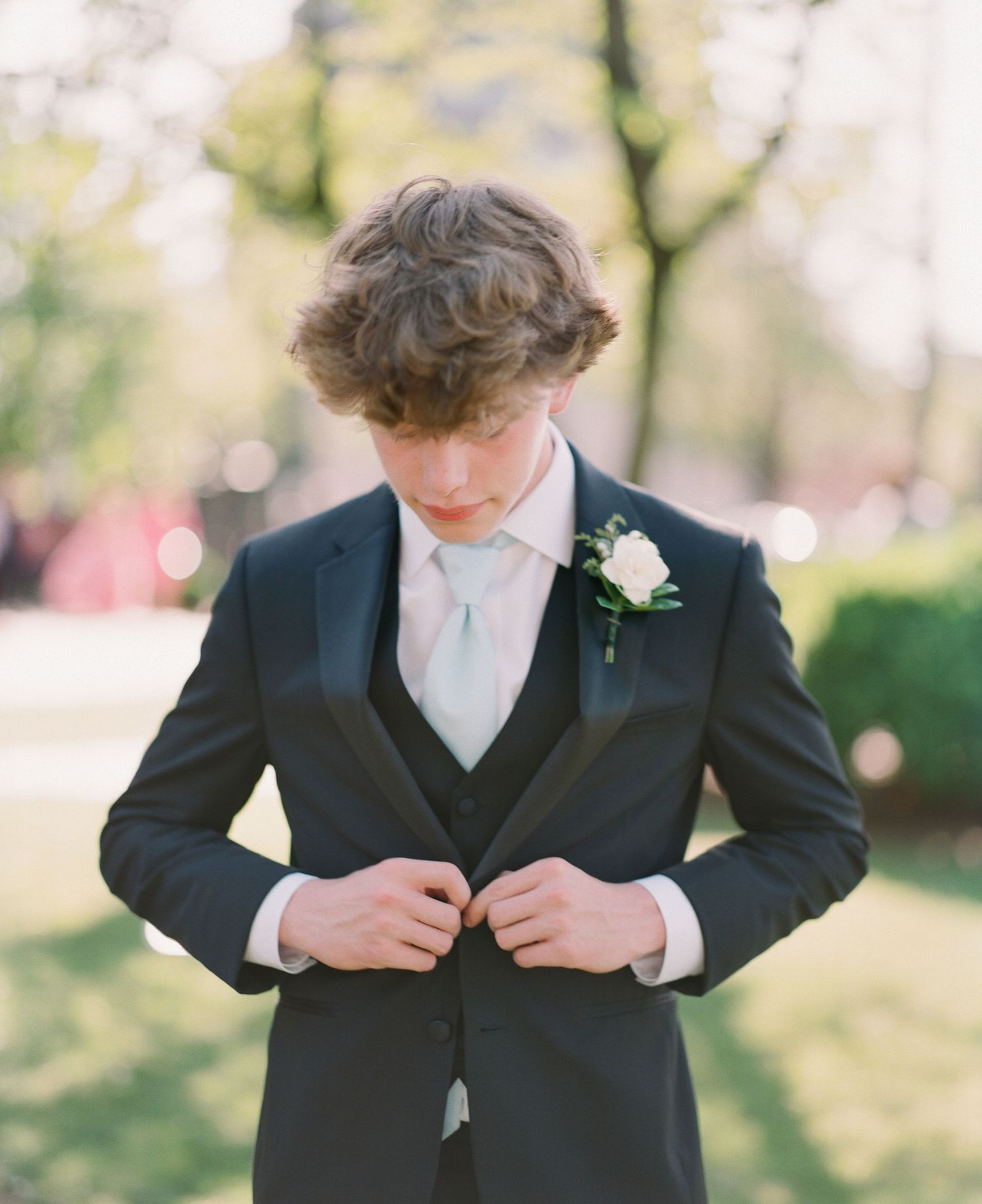 A young groom in a dark tuxedo with a white boutonniere adjusts his jacket buttons outdoors.