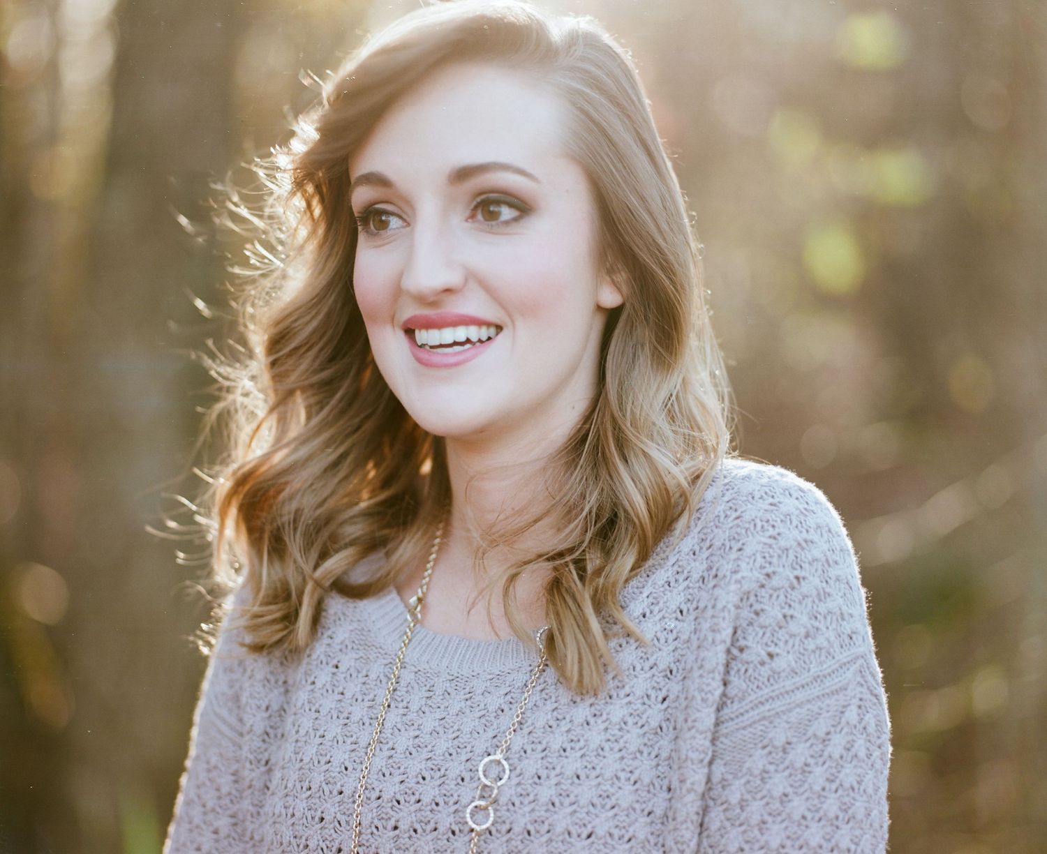 Woman in gray sweater smiling warmly in outdoor portrait with natural sunlit background.