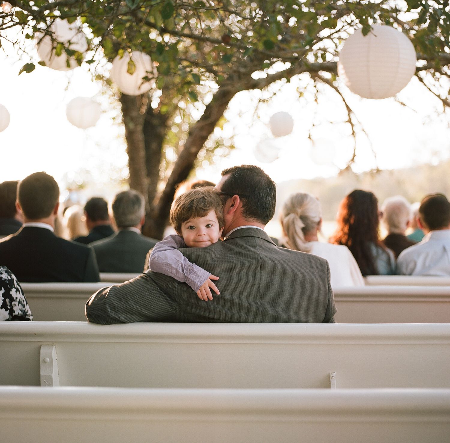 Wedding guests seated on white benches at an outdoor ceremony with hanging lantern decorations.