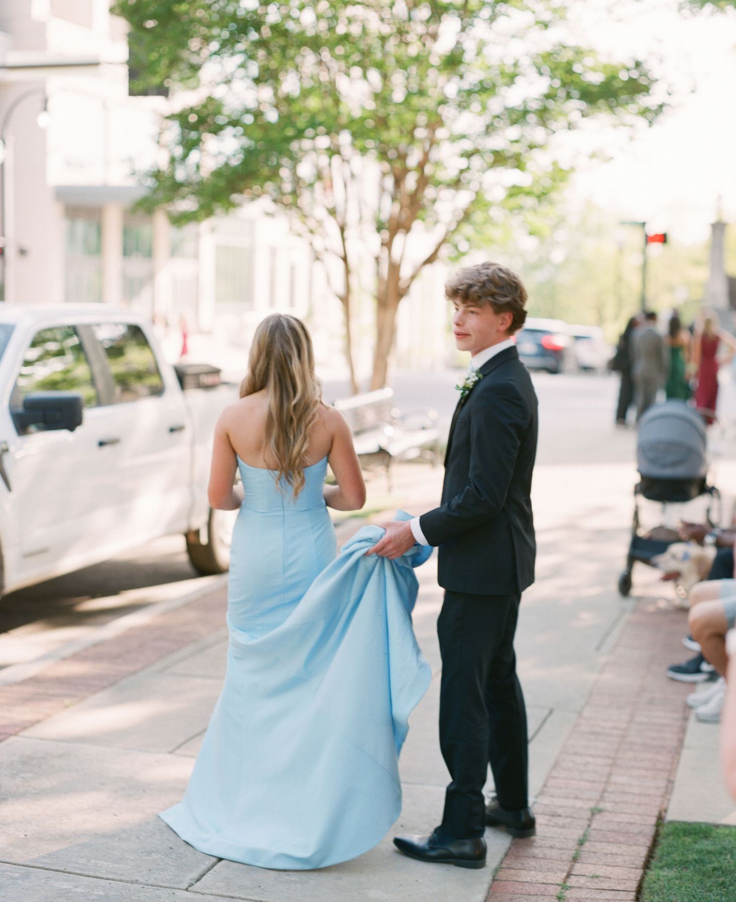 A young couple in formal attire walks on a sidewalk, the boy holding the girl's light blue gown train.