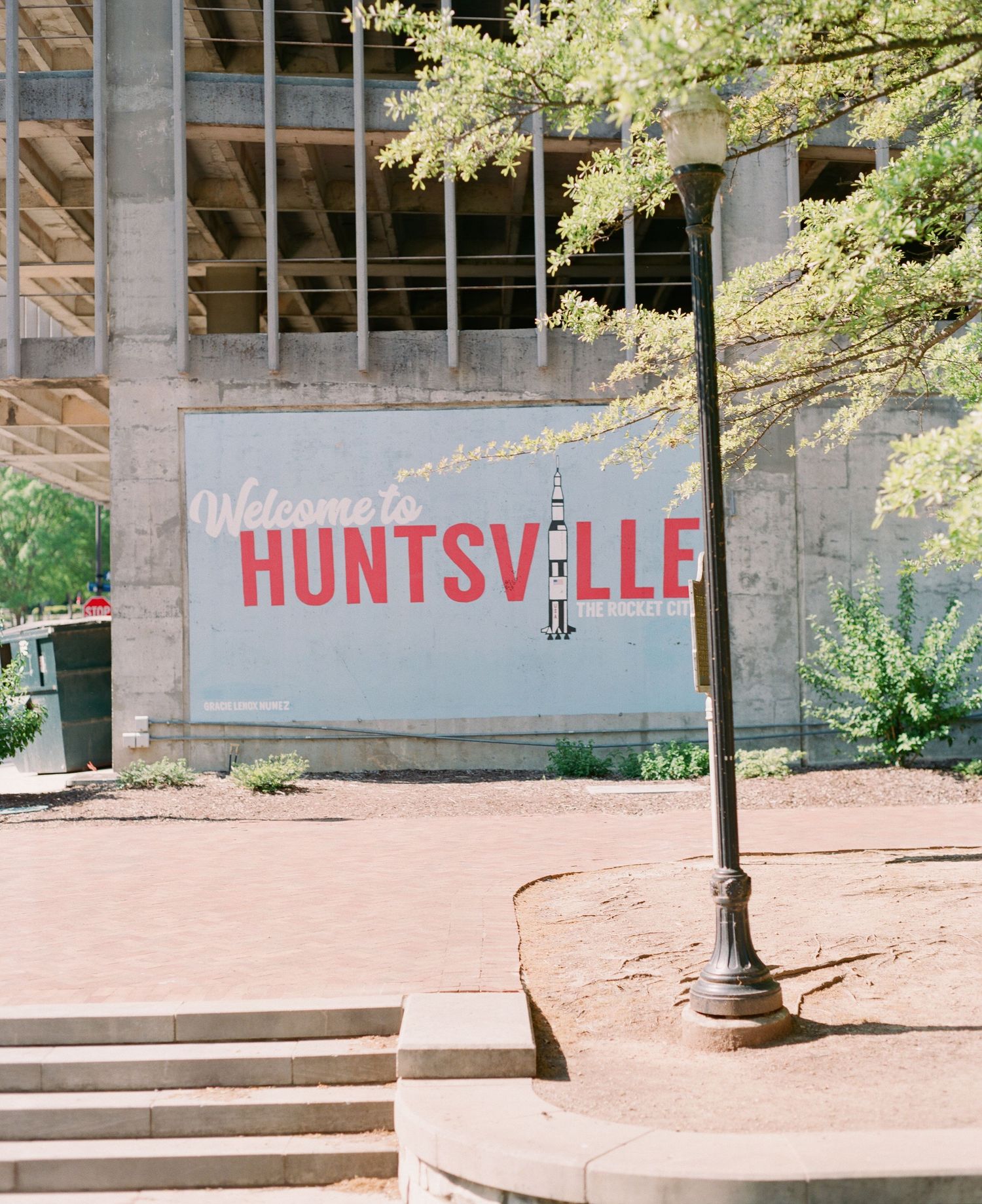 Welcome to Huntsville sign with rocket graphic displayed on a wall near a lamp post and green trees.