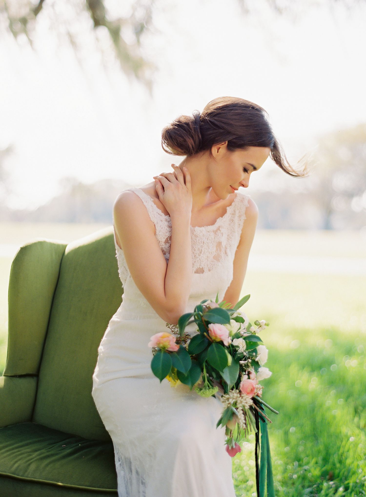 A bride in a white dress sits on a green vintage couch outdoors holding a floral bouquet with pink and green accents.