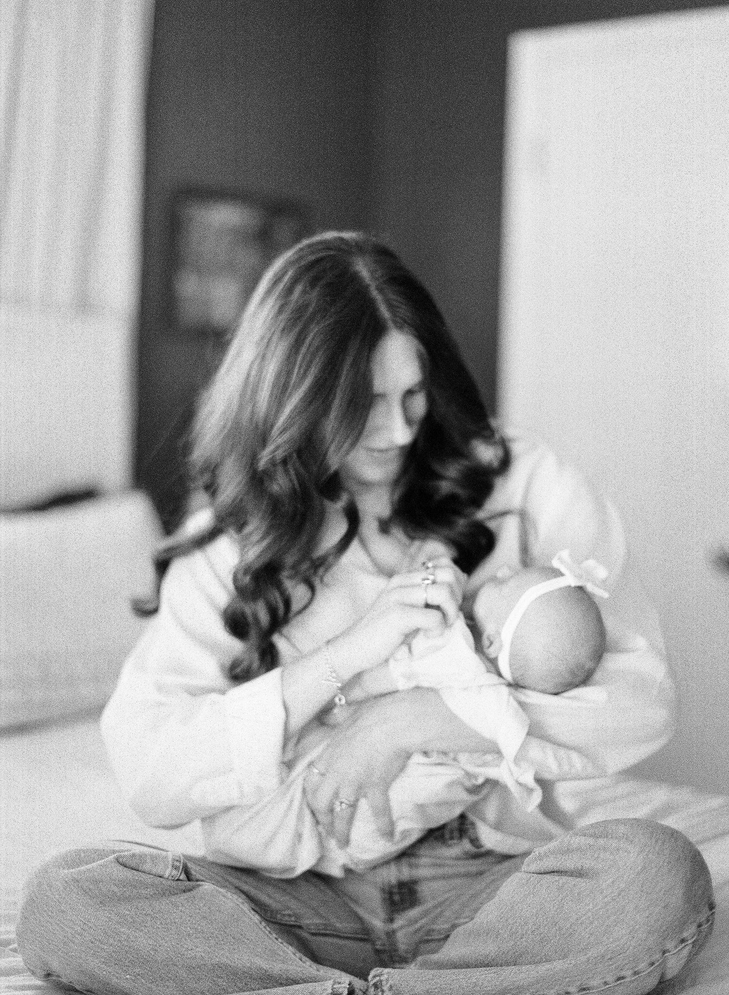 Black and white photo of a mother with long hair sitting cross-legged on a bed, tenderly cradling her newborn baby.