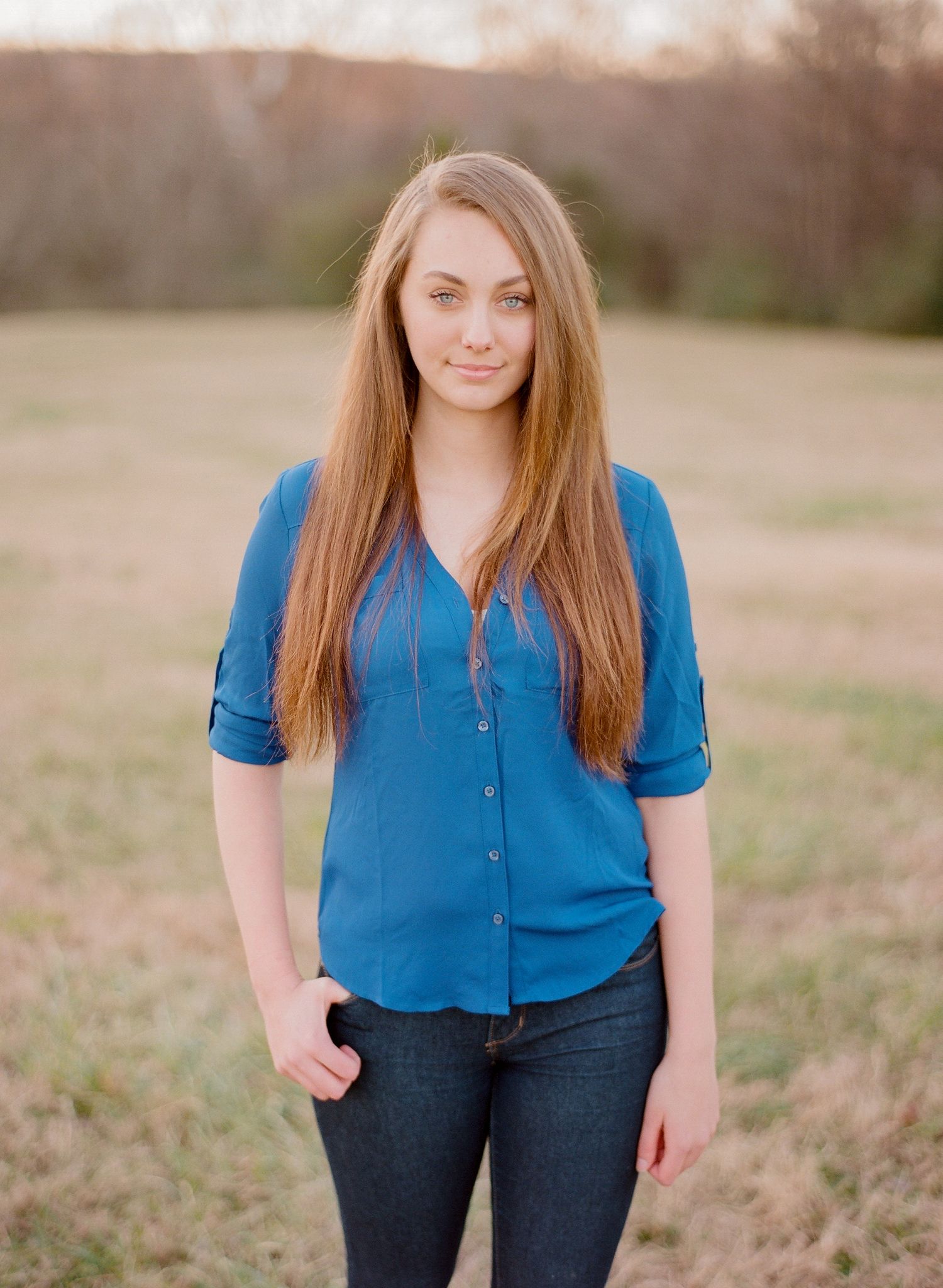 A woman in a bright blue blouse and dark jeans poses casually in a natural outdoor setting.