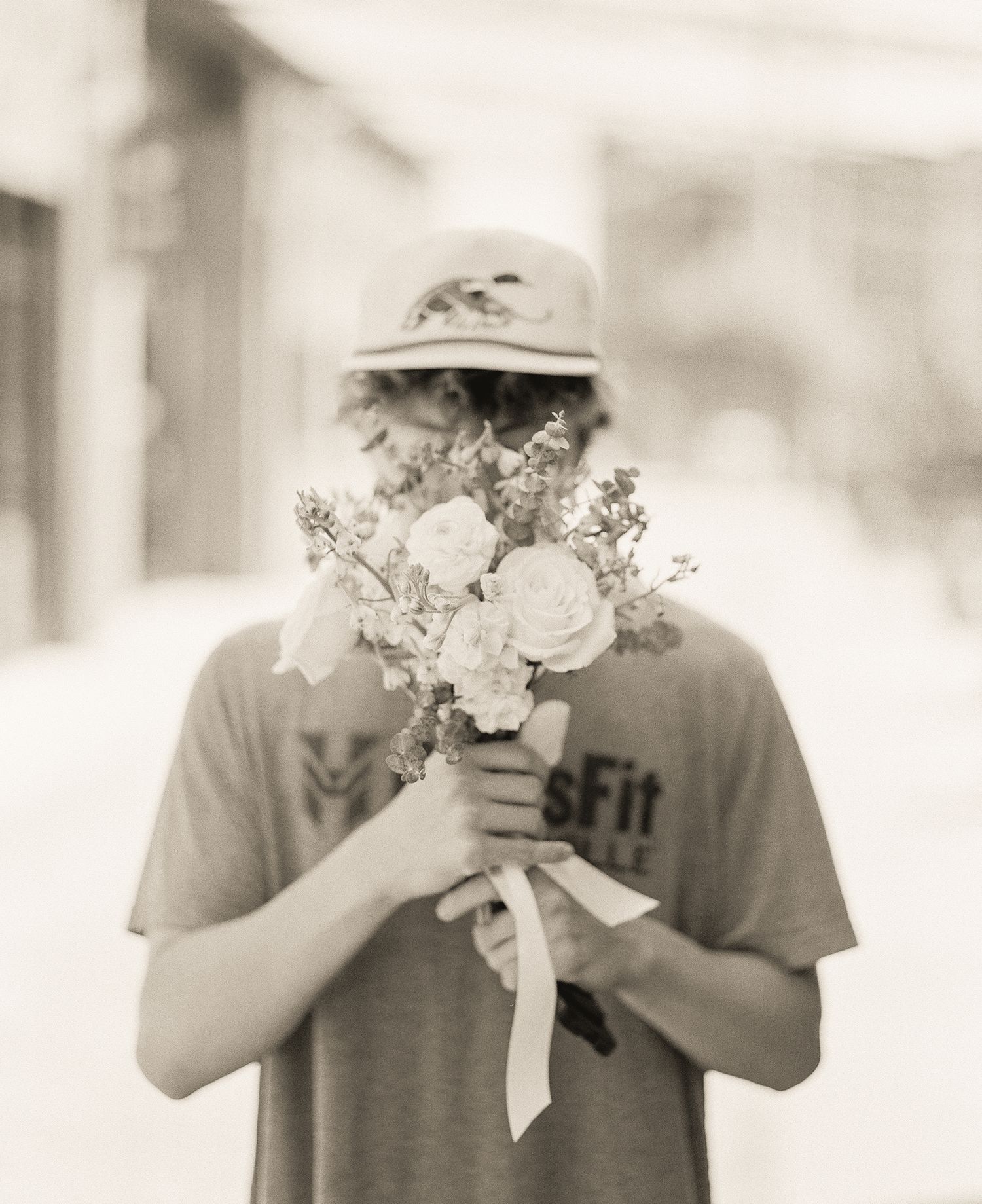 Person in bucket hat holding bouquet of white roses covering face on blurred street background.