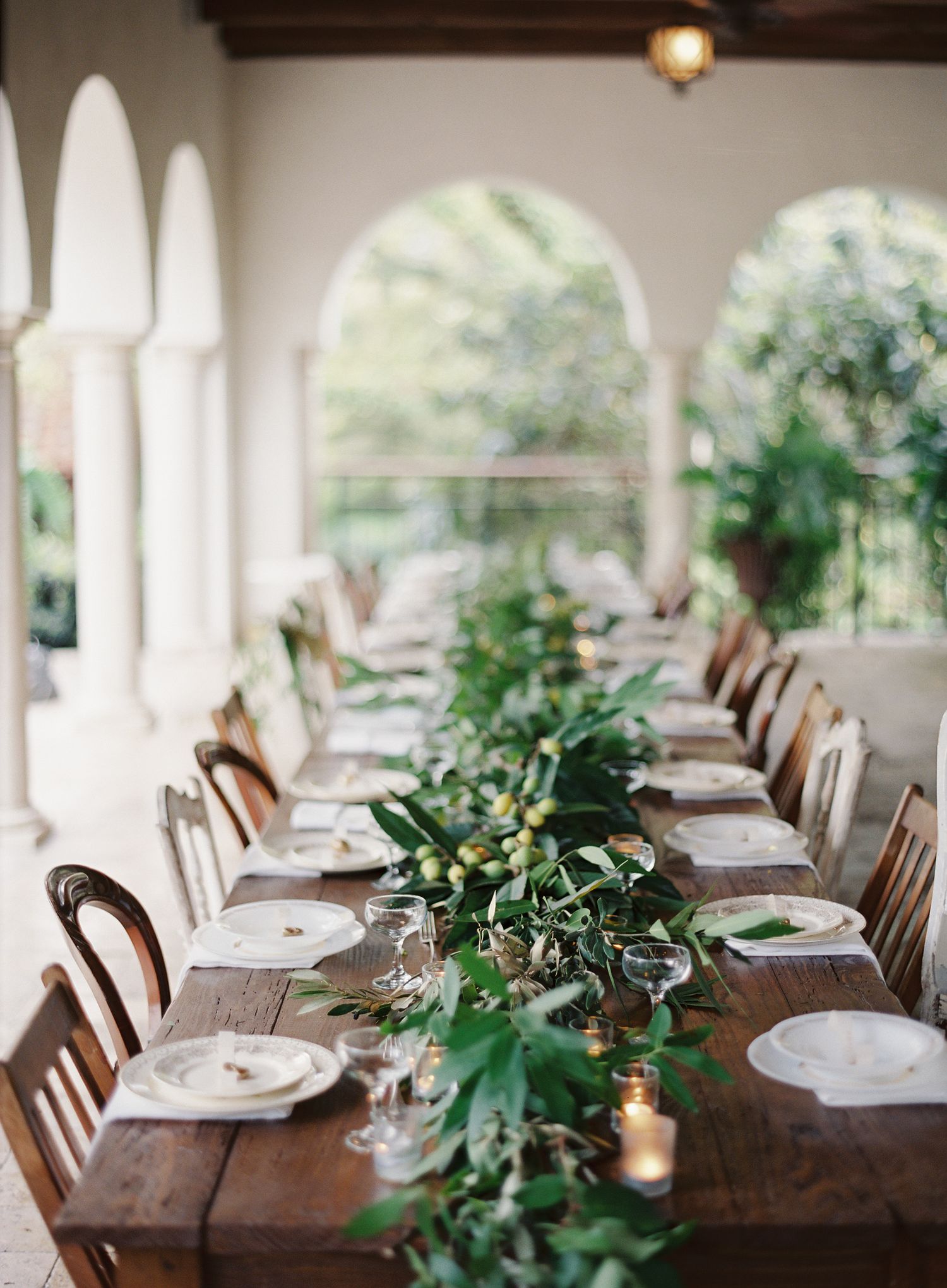Elegant rustic dining table set with greenery garlands and white place settings under arched colonnade.