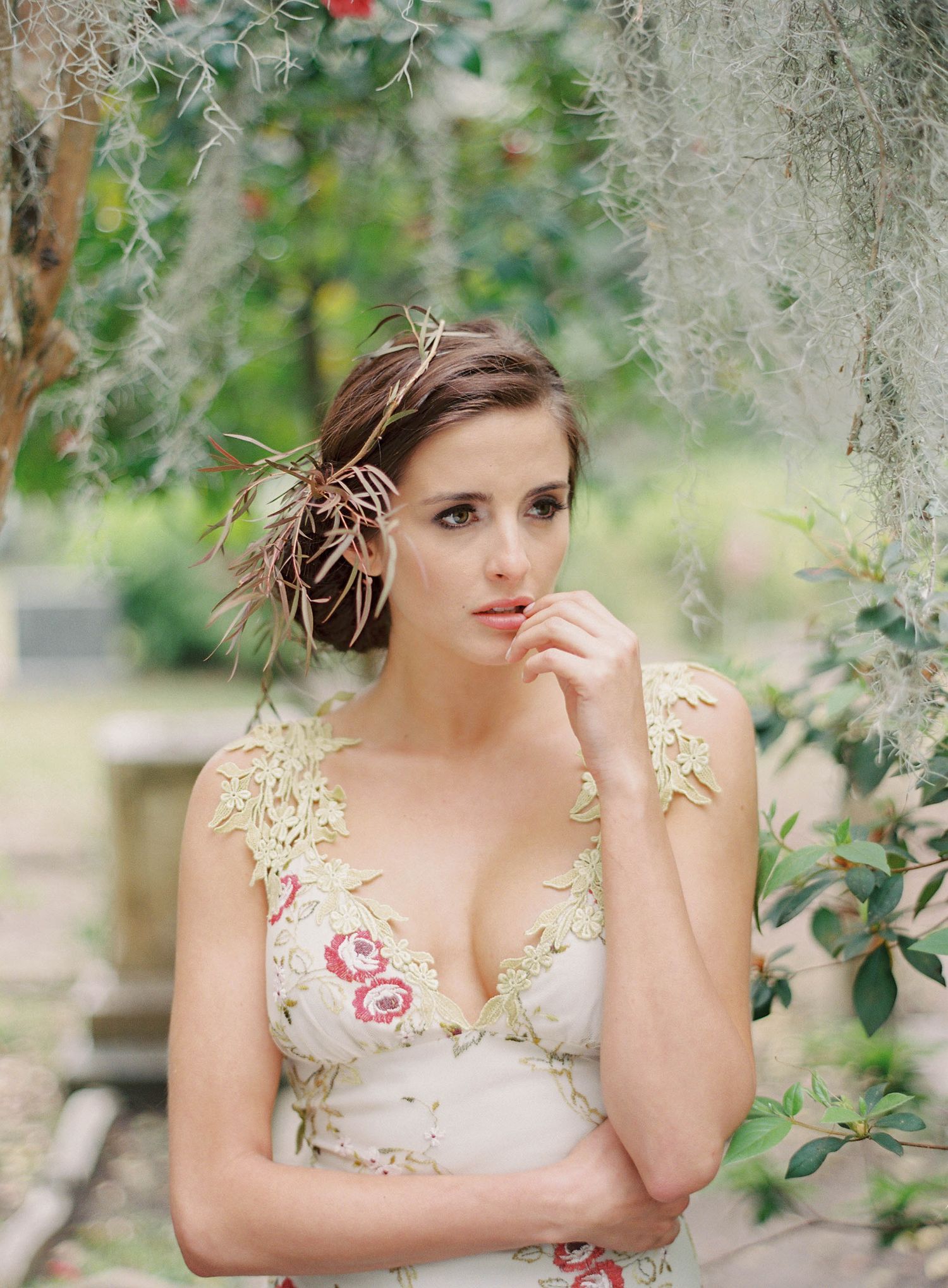 Bride in floral detailed dress poses among hanging Spanish moss and garden foliage.