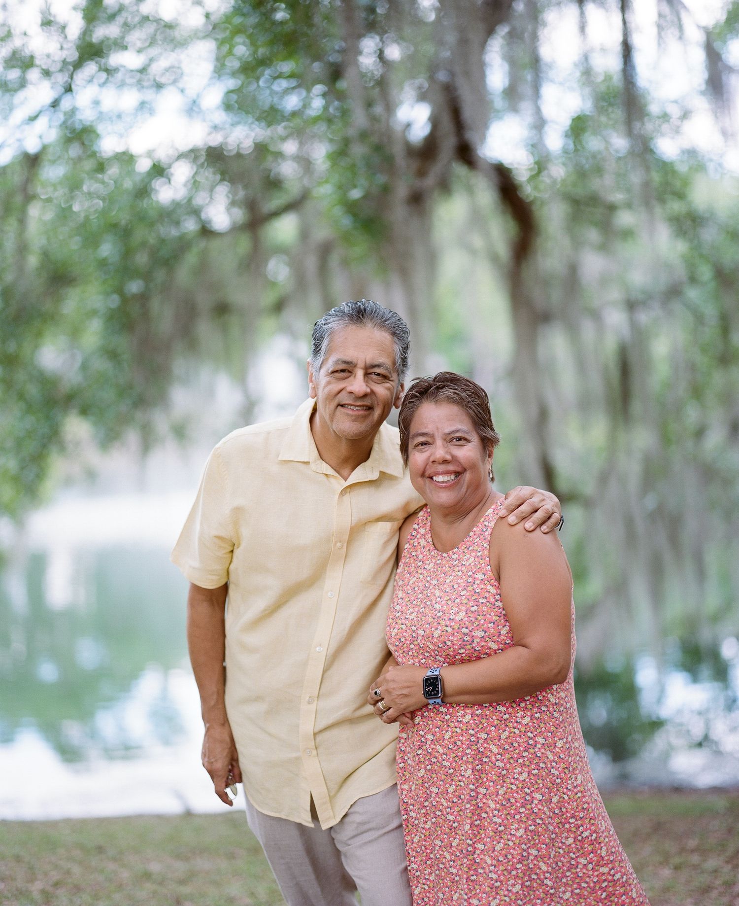 Couple in summer attire smiling together under Spanish moss trees near a lake.