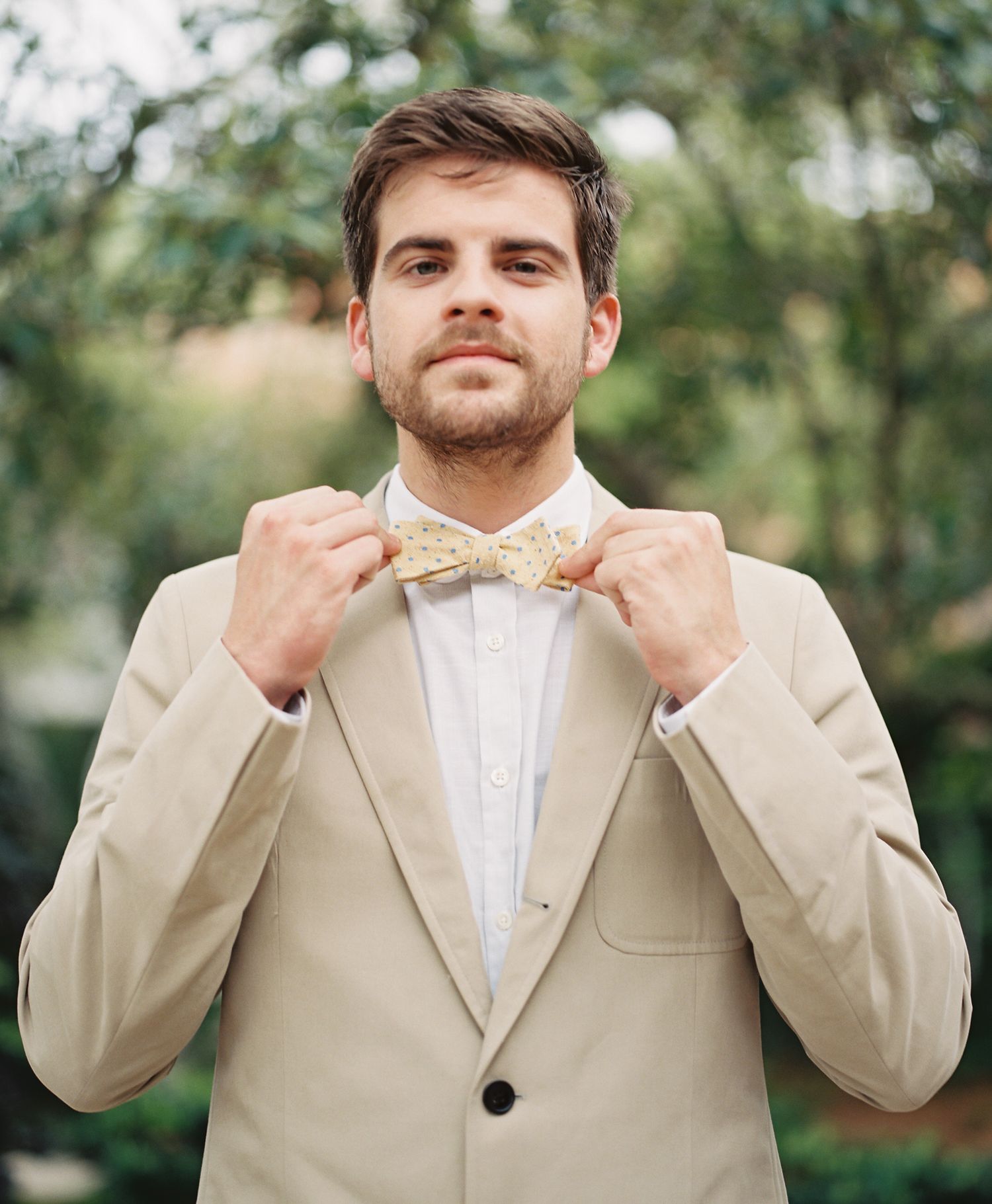 Close-up sequence of groom adjusting beige bowtie against natural outdoor backdrop.