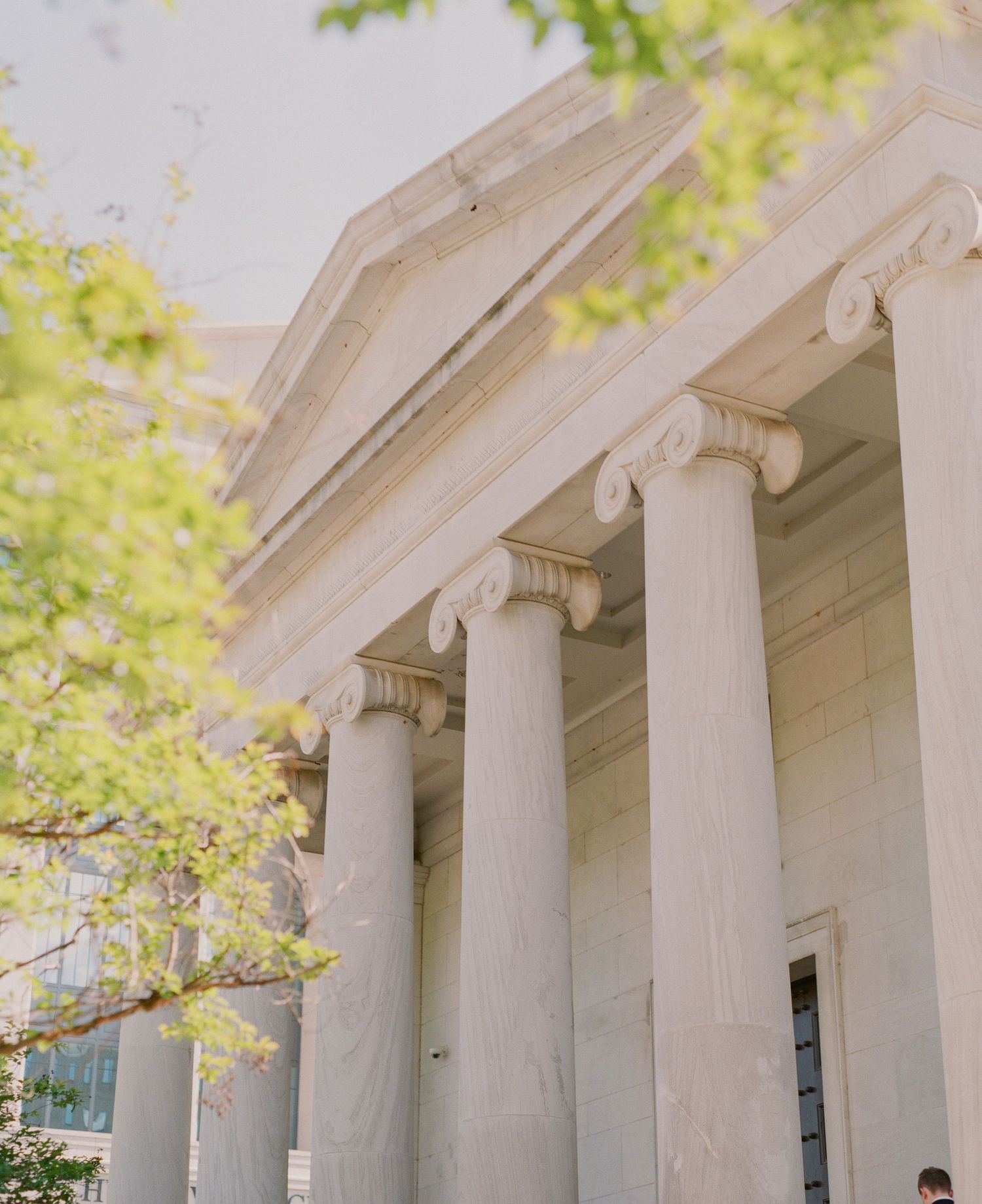 Neoclassical building with tall Ionic columns and white stone facade surrounded by lush green trees.