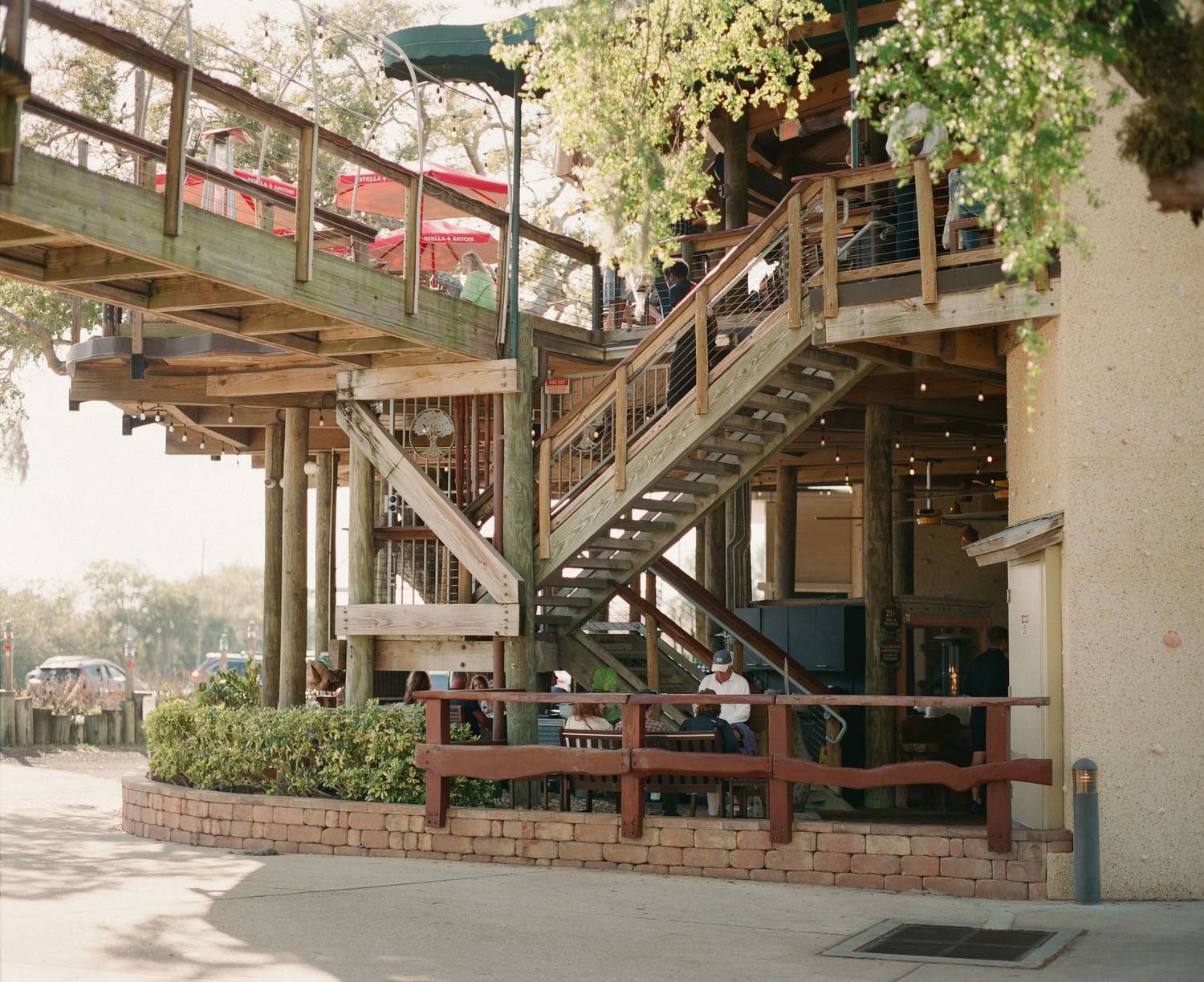 Multi-level wooden deck restaurant with staircases, brick planters, and lush trees in an outdoor dining setting.