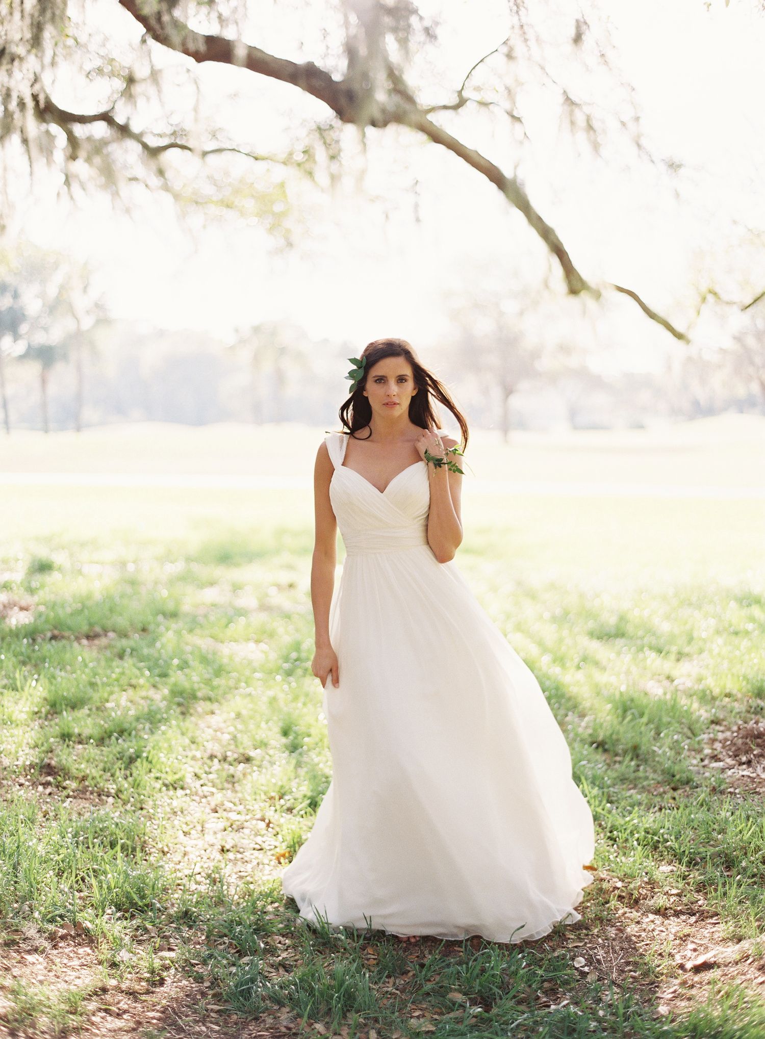A bride in a flowing white wedding dress poses in a sunlit outdoor setting with trees in the background.