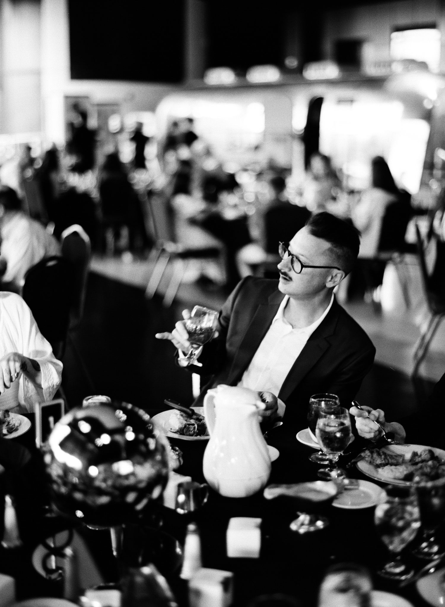 Man in black suit with glasses sits at elegant dinner table in black and white event photography.