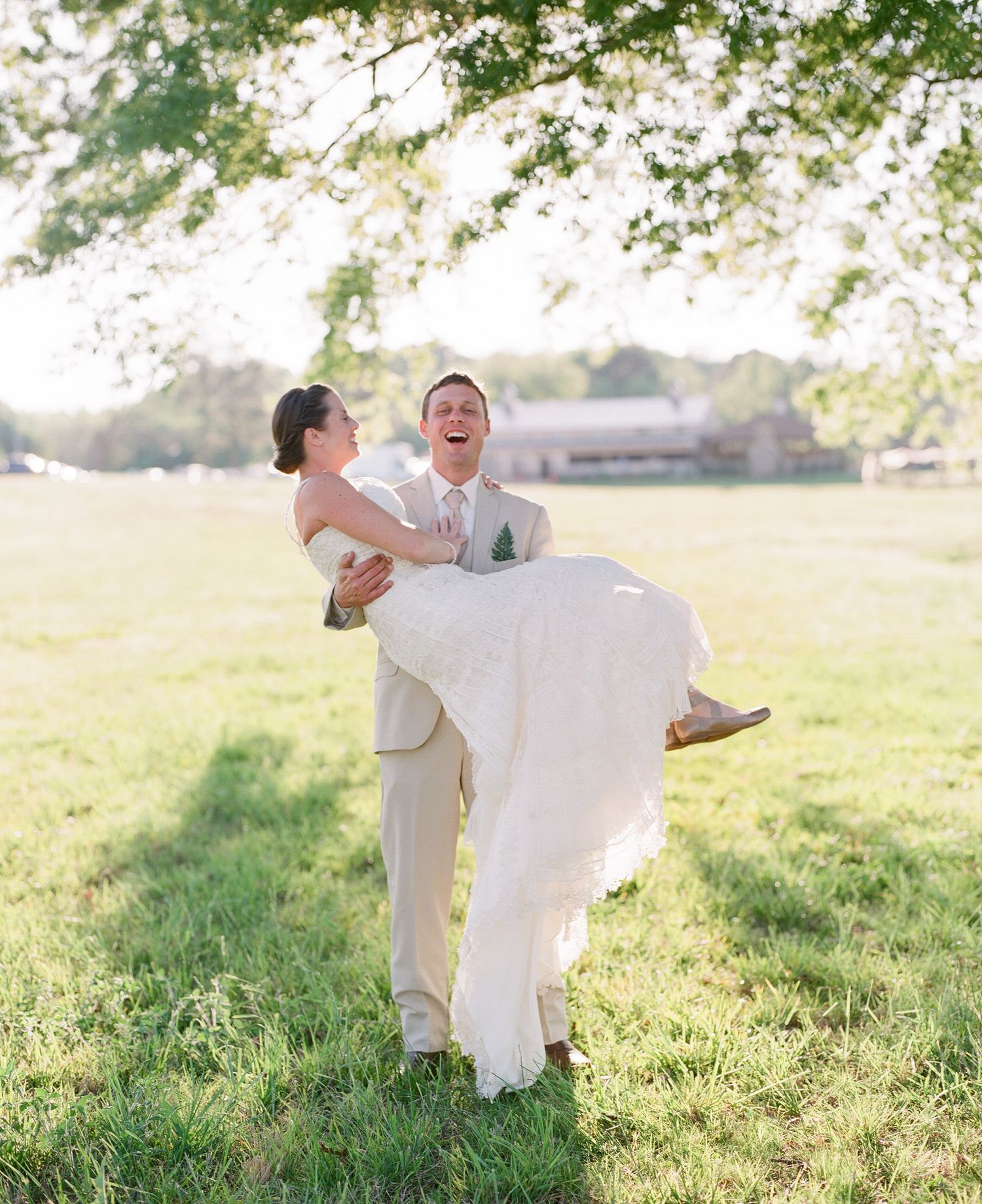 Groom carries bride in romantic gesture across sunny meadow in summer wedding scene.