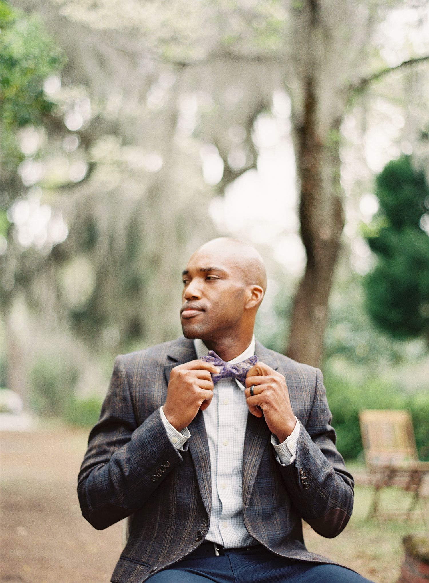 Man adjusting his bowtie while seated outdoors among trees.