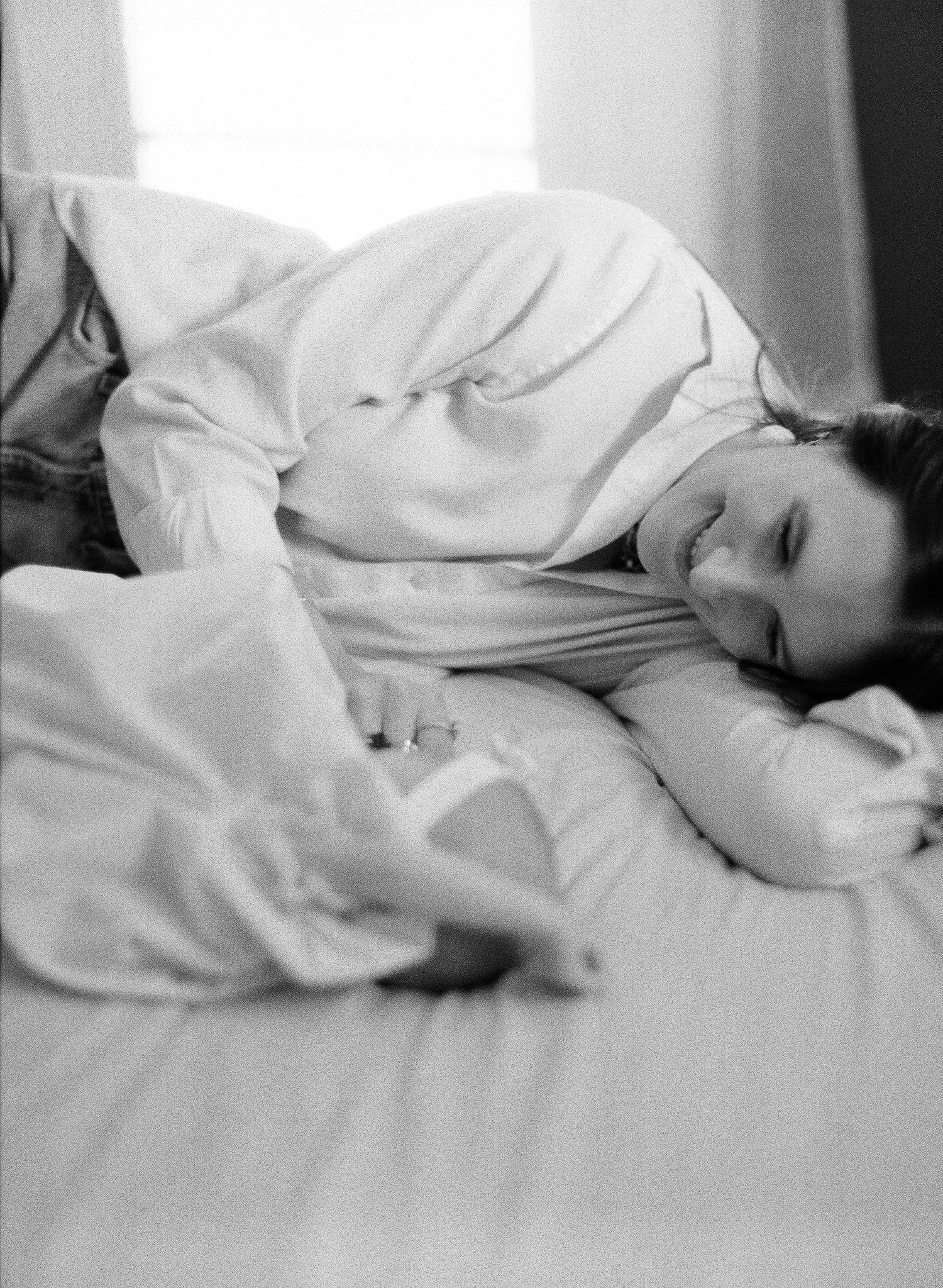 Black and white photo of a woman lying on a bed, smiling, wearing a white shirt, resting her head on a pillow.